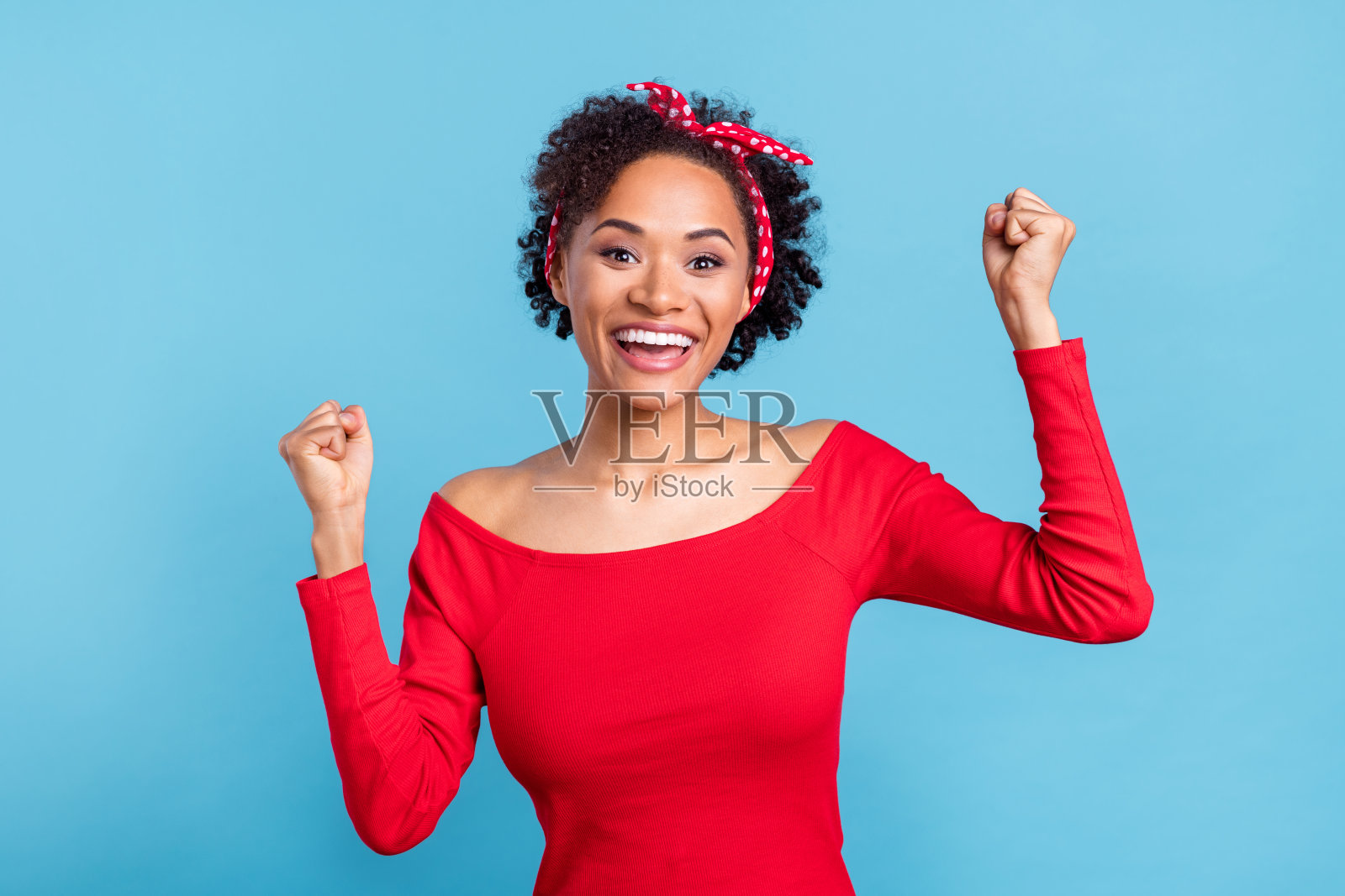 一位穿着红色露肩衬衫的美丽非洲女士,微笑着举起双拳,背景为蓝色。照片摄影图片
