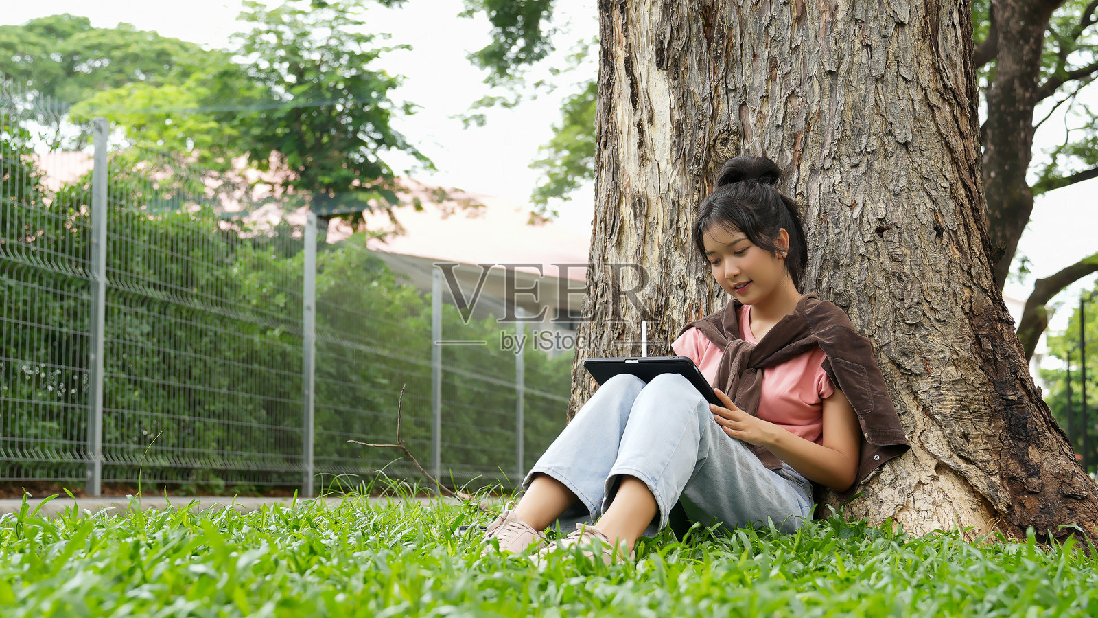 一名高中生靠在树上,专注于在平板电脑上写作。学校的红砖外墙在背景中可见。照片摄影图片