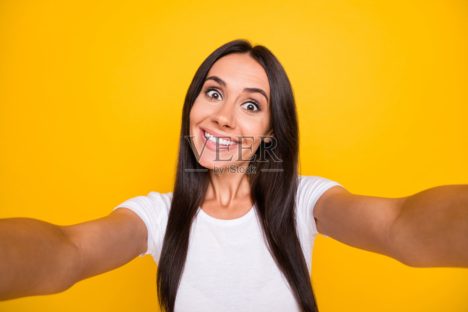 一位穿着白色T恤的年轻女性在黄色背景下微笑着拍摄自拍照,显得光鲜有趣。照片摄影图片