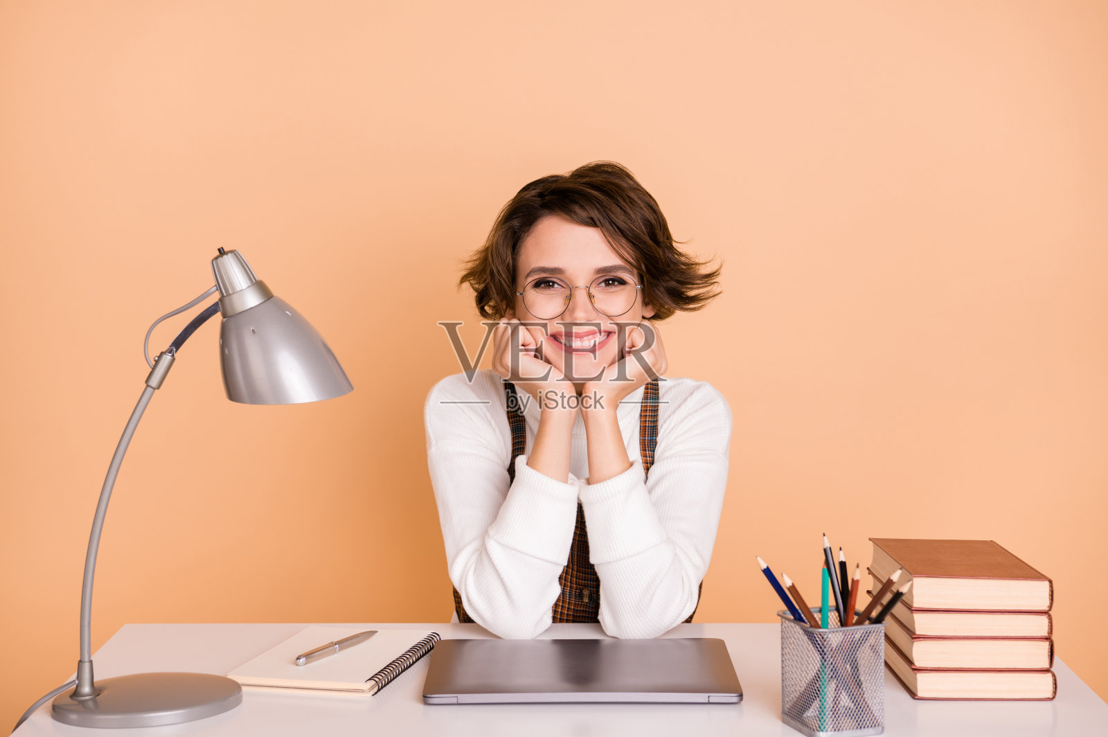 乐观可爱的短发女孩坐着,双手托着脸,穿着毛衣,背景为桃色。照片摄影图片