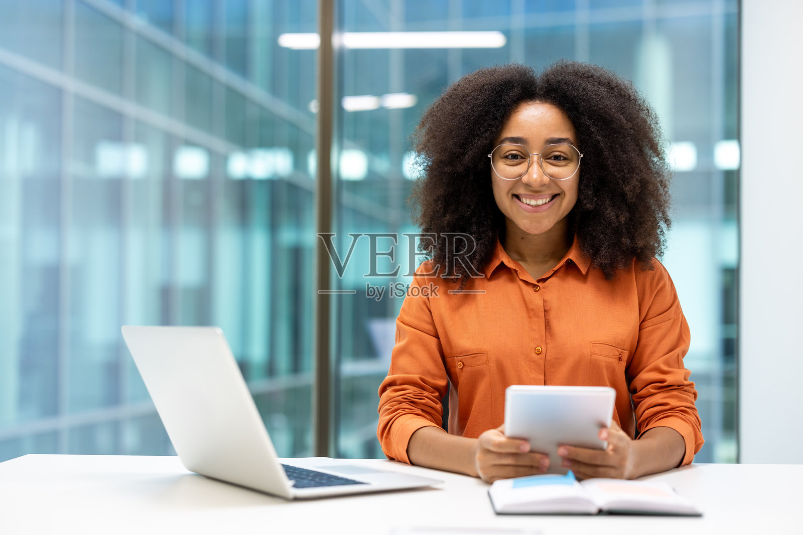 微笑的戴眼镜女性在现代办公室环境中手持平板电脑,旁边有一台笔记本电脑。照片摄影图片