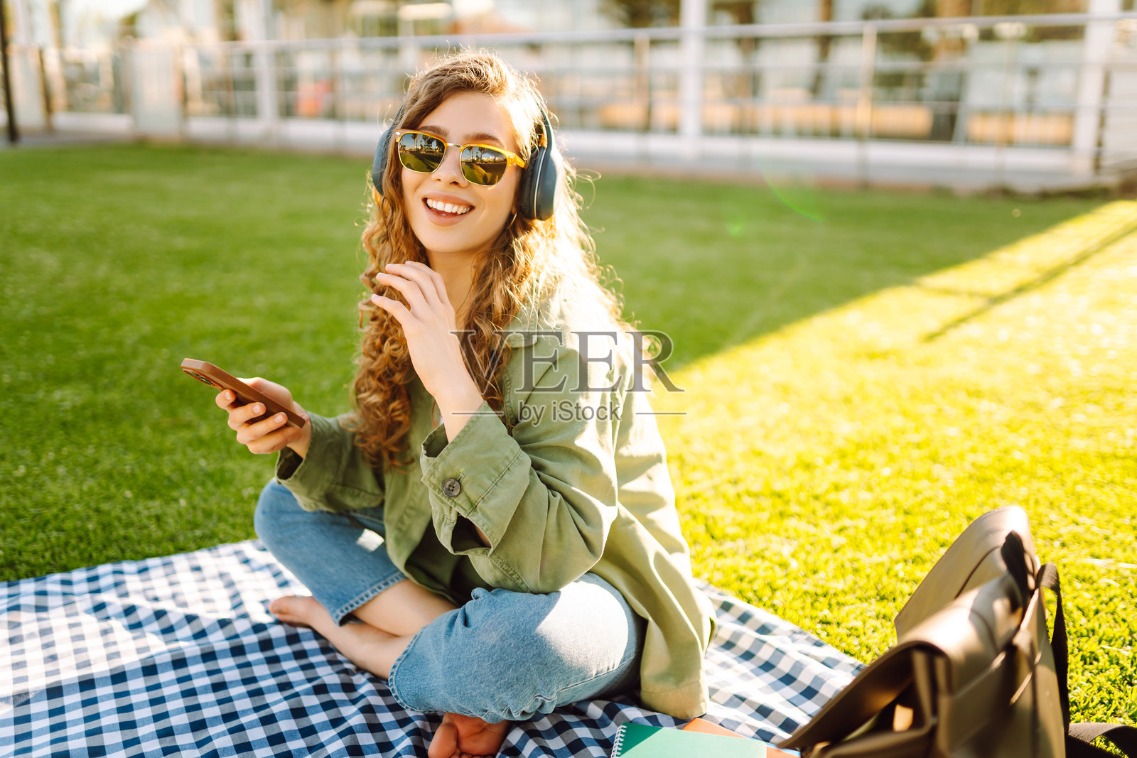 年轻女性穿着休闲服,戴着无线耳机,使用手机,坐在草坪上的垫子上,周围是棕榈树。照片摄影图片