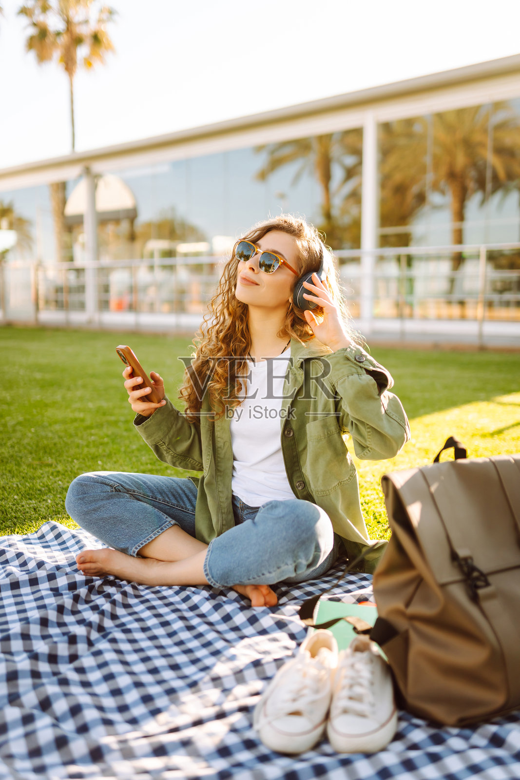年轻女性穿着休闲服,戴着无线耳机,使用手机,坐在草坪上的垫子上,周围是棕榈树。照片摄影图片