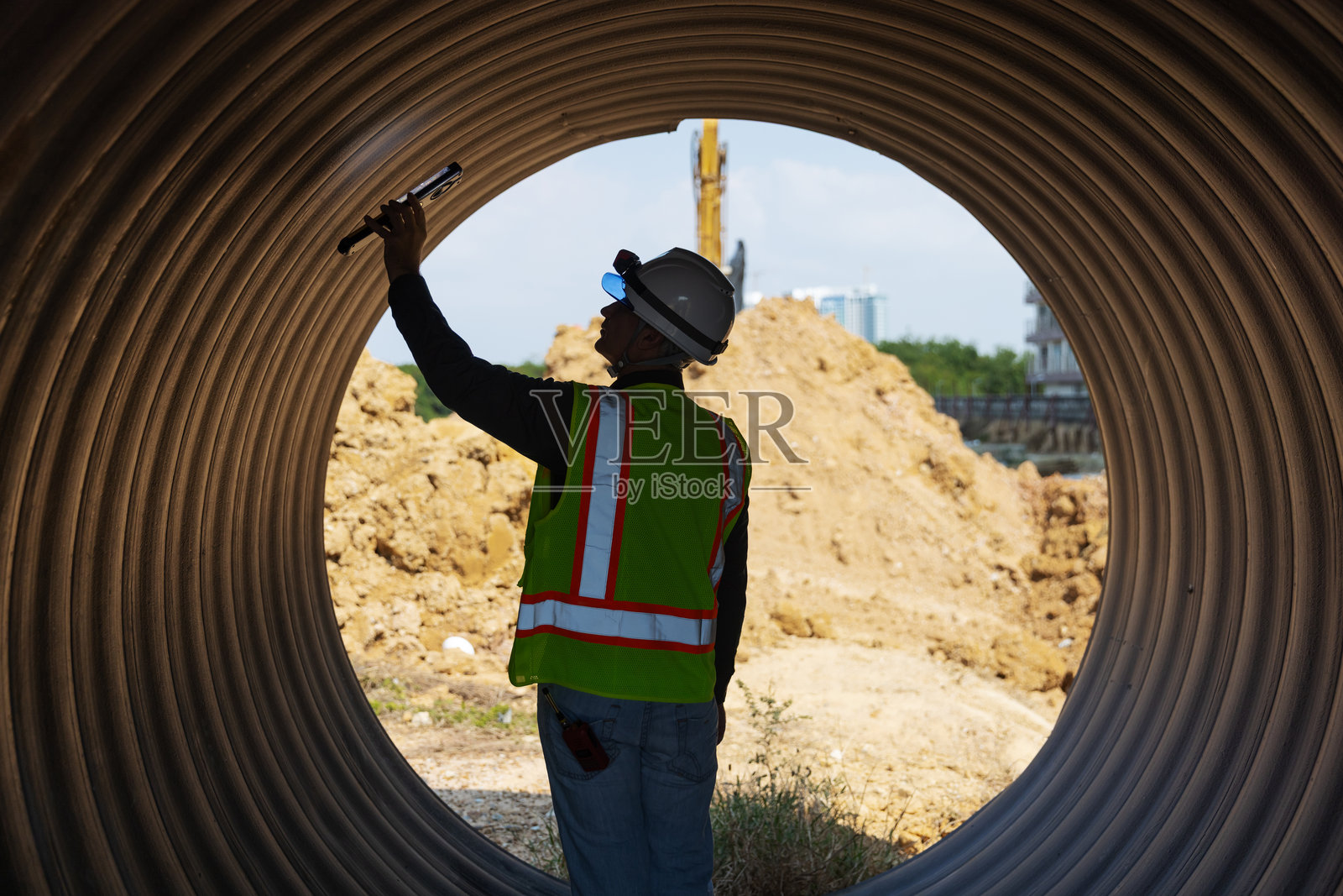 一名穿着安全装备的建筑工人在用手电筒检查大型波纹排水管的内部。照片摄影图片