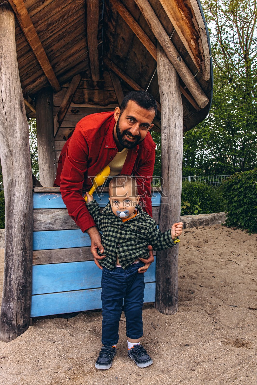 父亲和幼儿在户外游乐场的木制游乐屋里一起玩耍。孩子用一个黄色的铲子倒沙子,周围是自然的木质纹理和绿色植物。照片摄影图片