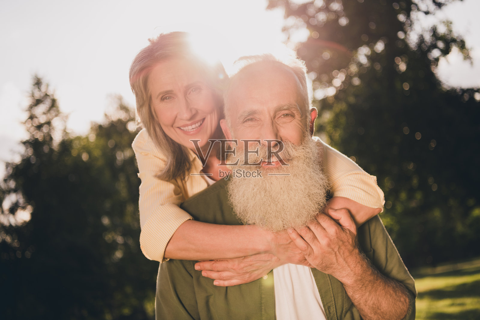 乐观的白发老年夫妇在公园外穿着休闲衬衫,互相拥抱,骑在对方背上散步。照片摄影图片