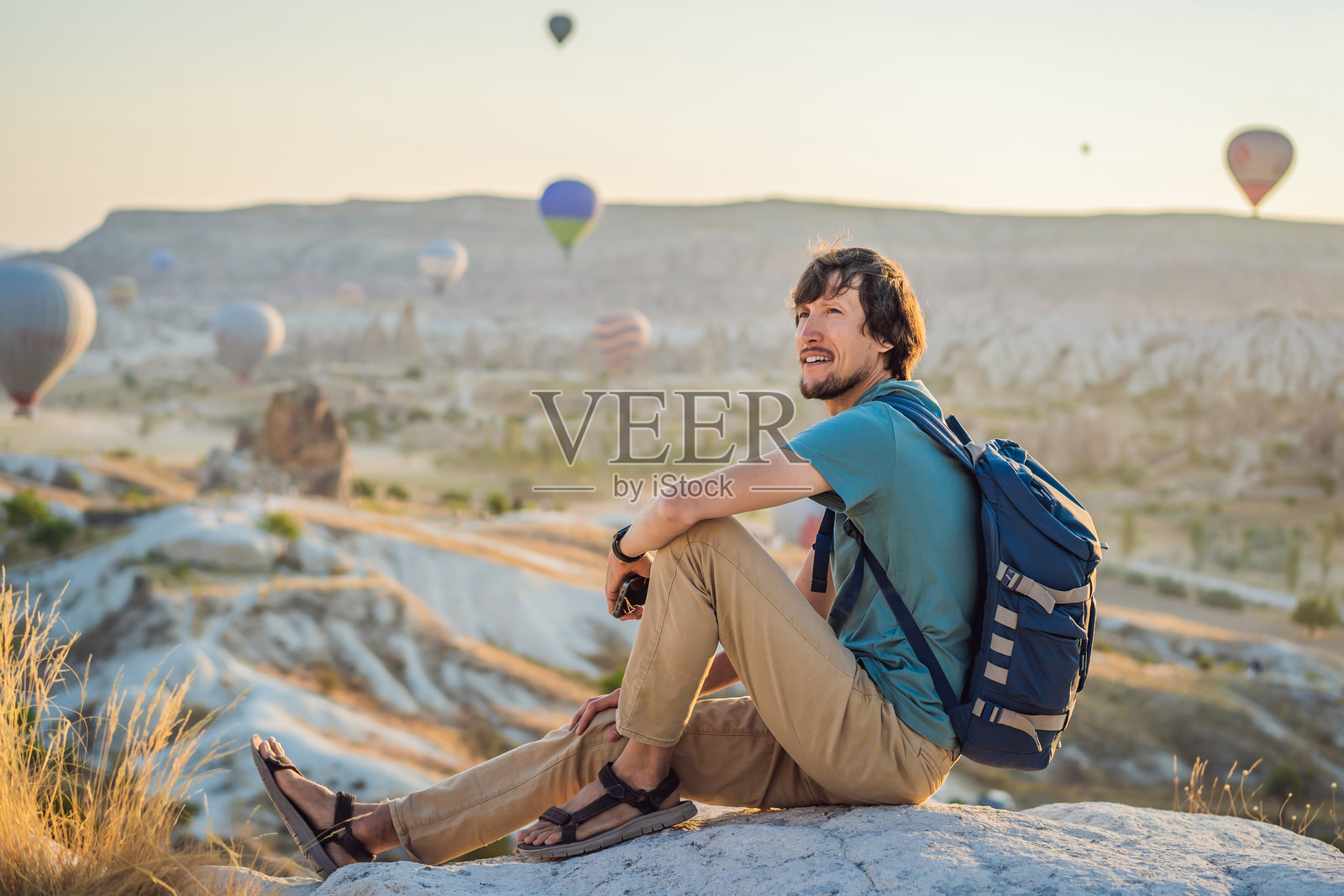 在土耳其卡帕多奇亚,一名游客在欣赏热气球。快乐的土耳其旅行概念。男子在山顶享受美妙的景色。照片摄影图片