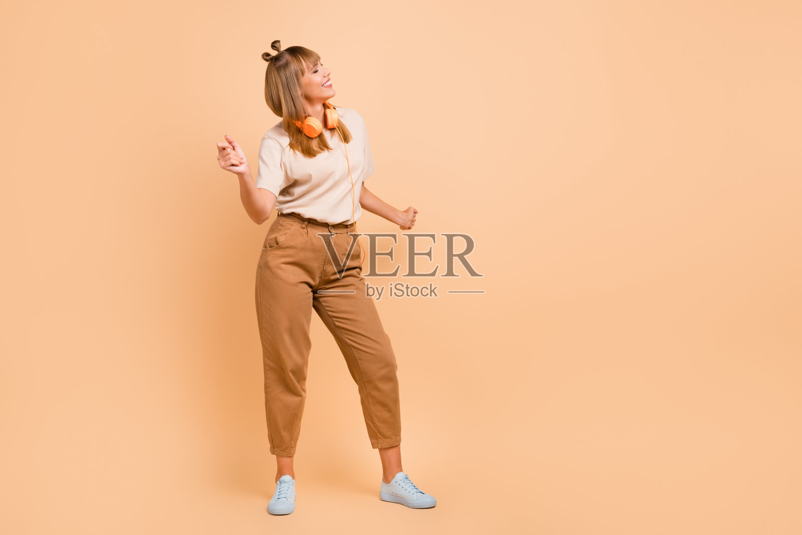 可爱女孩全身照,闭眼享受舞蹈,戴着耳机,穿着T恤,背景为米色。照片摄影图片