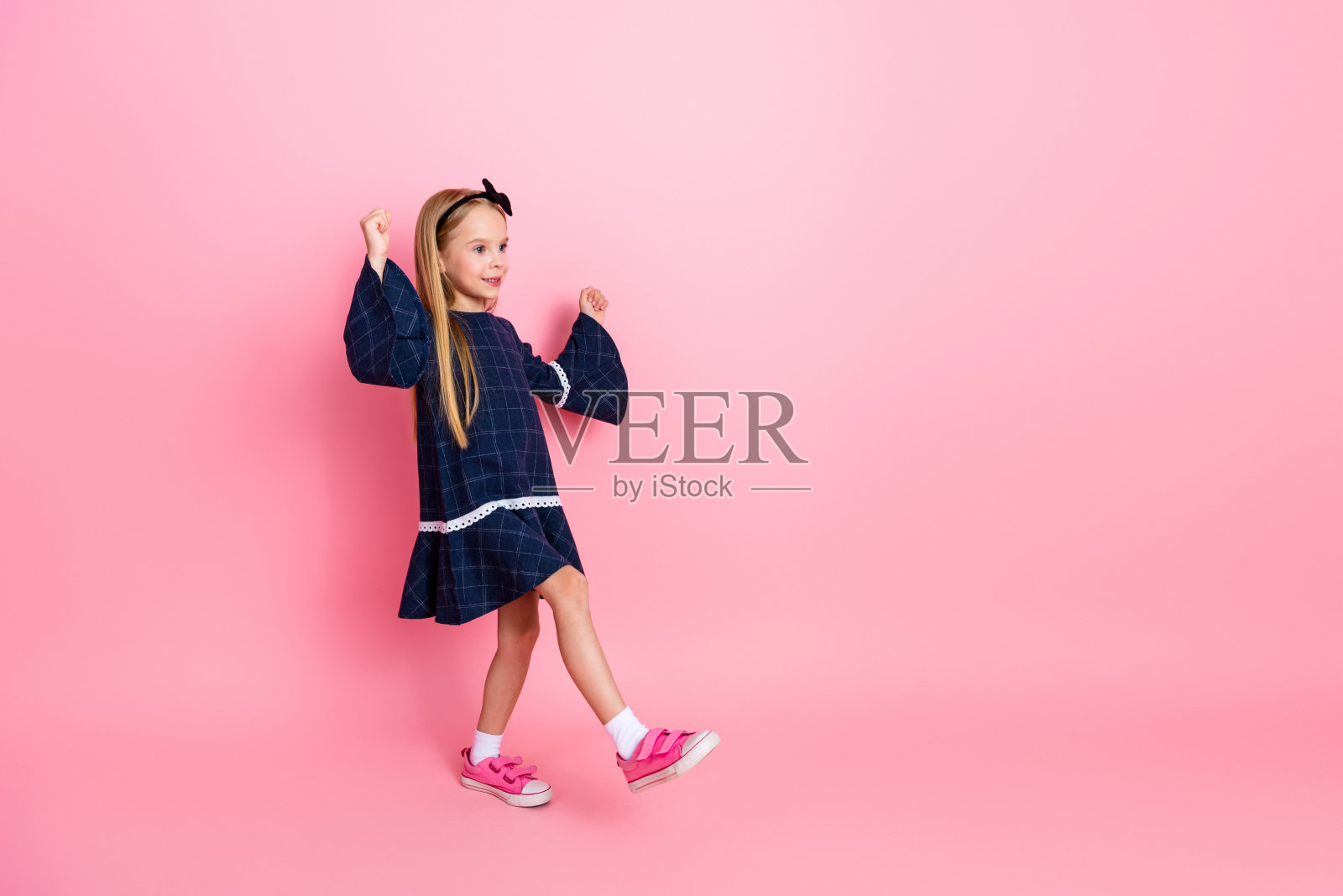 可爱的小女孩穿着深蓝色裙子和粉色鞋子,在明亮的粉色背景前快乐地摆姿势,面带愉悦的表情。照片摄影图片