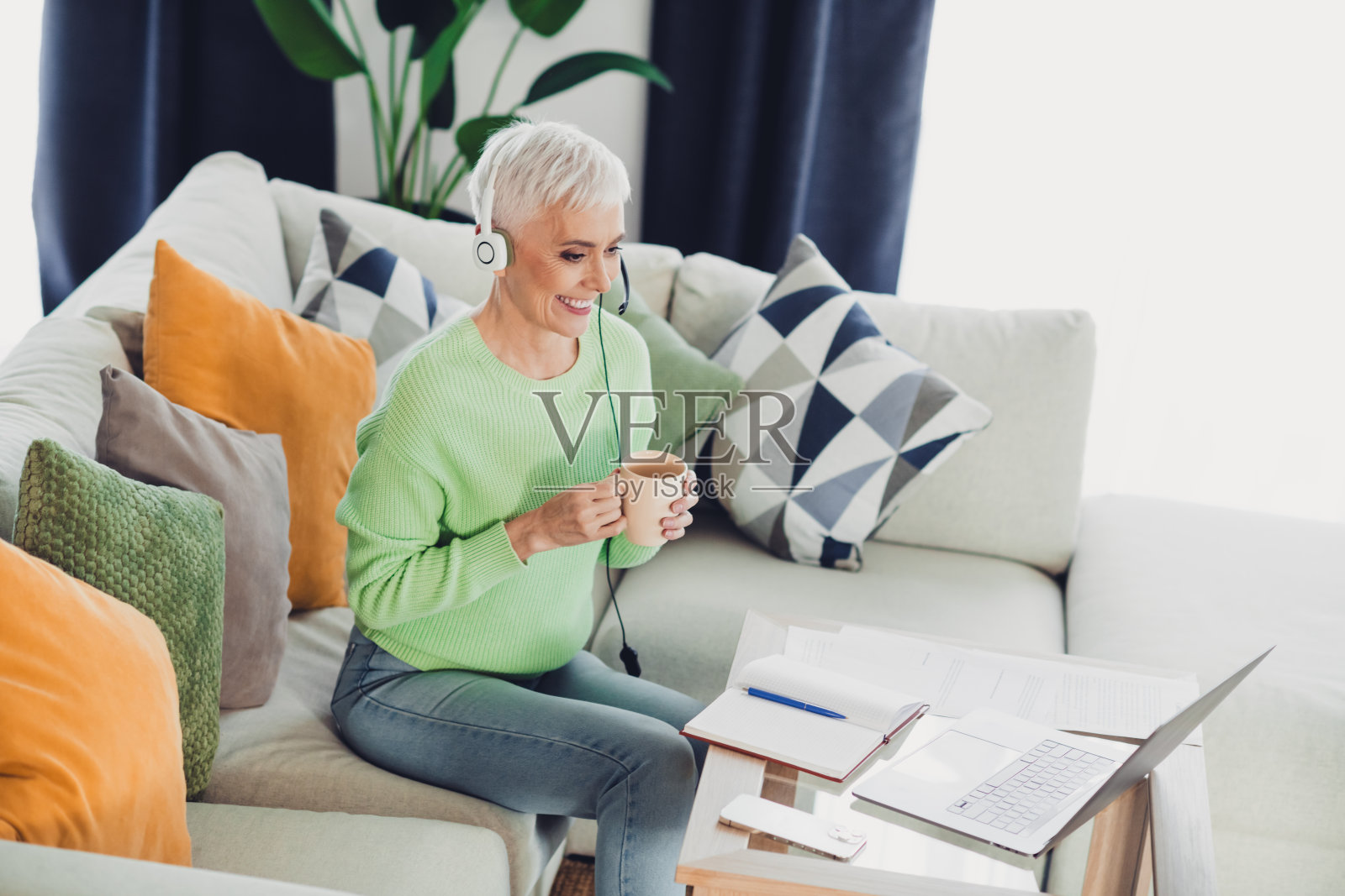 成熟的女性在家中享受轻松的视频通话,佩戴耳机,手握咖啡杯,体现了休闲与舒适。照片摄影图片