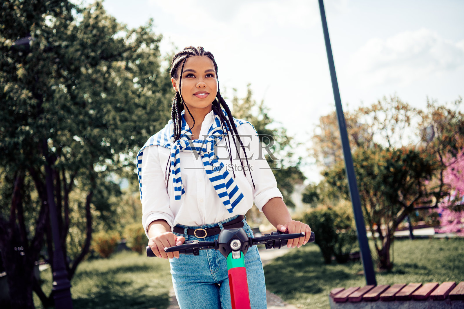 可爱的女性扎着辫子,在阳光明媚的户外骑着滑板车,享受美丽的城市花园。照片摄影图片