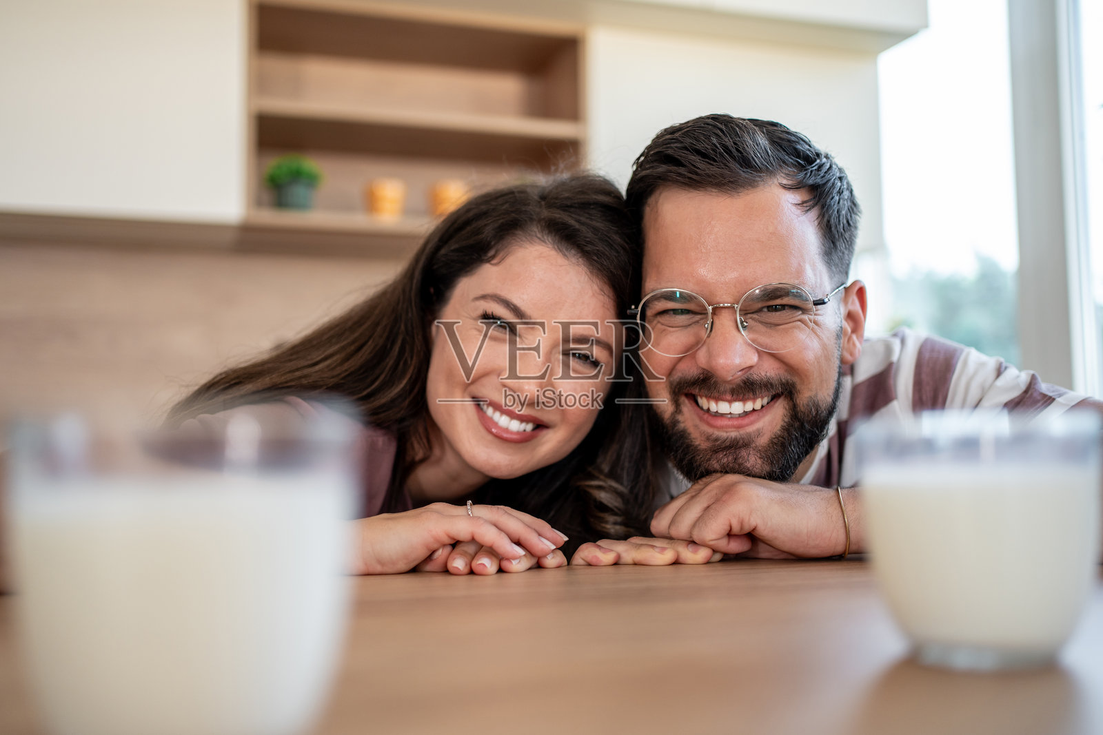 快乐的情侣在厨房里微笑着对着镜头,手里拿着牛奶杯。照片摄影图片