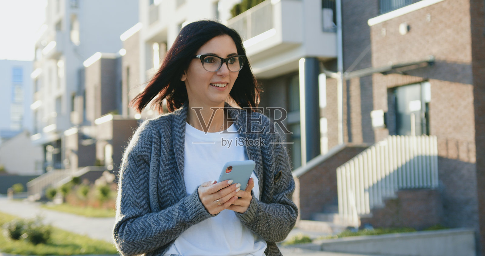 年轻美丽的白人自由职业女性在阳光明媚、微风拂面的城市街道上走路时使用智能手机照片摄影图片