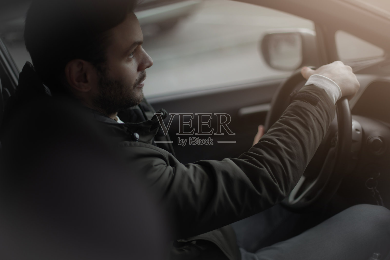 年轻男子侧面驾驶汽车,穿着深色夹克,专注于道路。照片捕捉了一个平静而专注的驾驶时刻,柔和的自然光线洒在车内。照片摄影图片