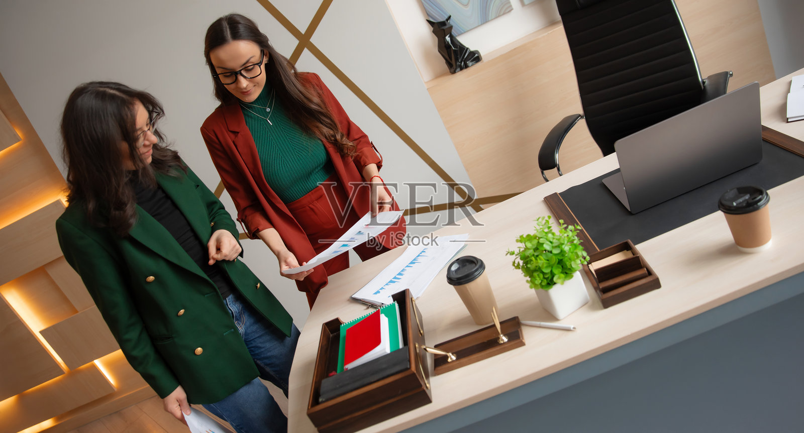 两位女商人在现代办公室桌前查看图表,桌上有笔记本电脑、咖啡杯和绿色植物。这个时尚的工作空间拥有温暖的灯光、简洁的线条和优雅的布局。照片摄影图片