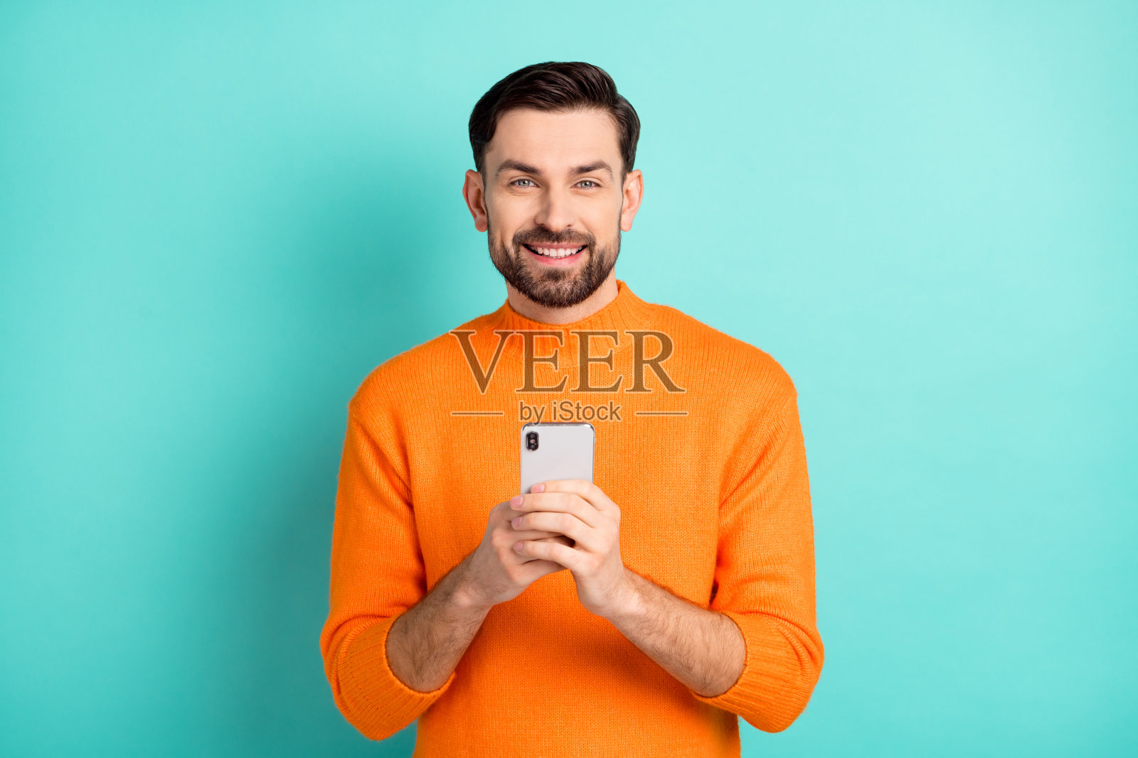 年轻快乐的男子面带微笑,愉快地使用手机聊天,背景为青色。照片摄影图片