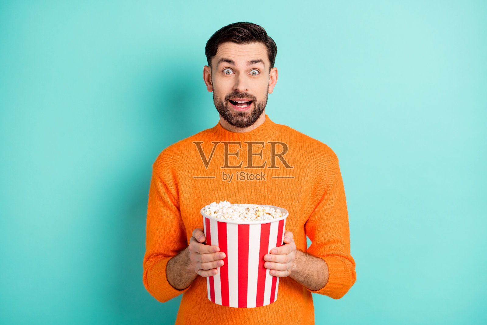 年轻兴奋的男子惊讶地拿着爆米花盒子观看电影,背景为青绿色。照片摄影图片