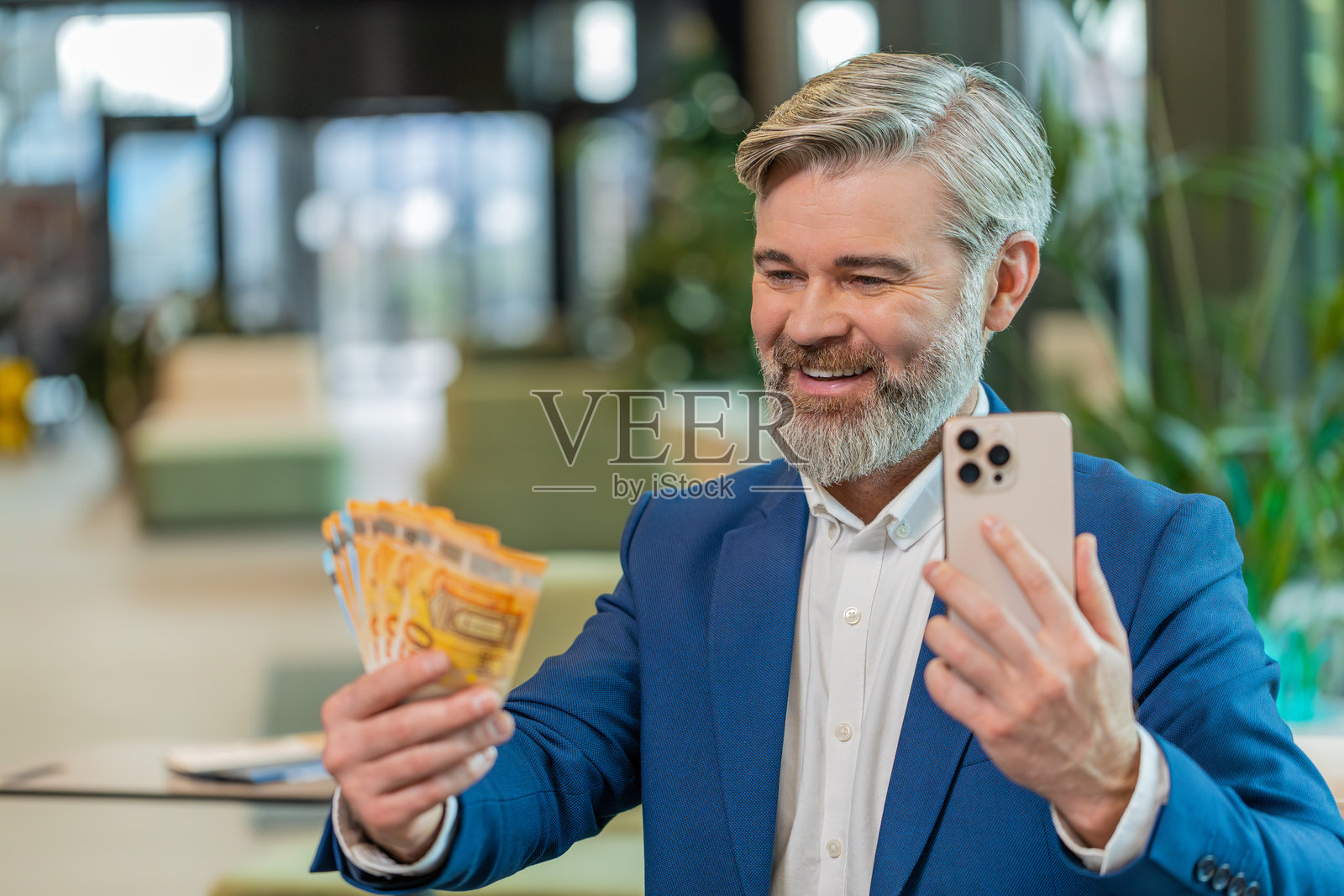 成功的商人坐在办公室大堂,手持智能手机,数着一叠欧元钞票。照片摄影图片