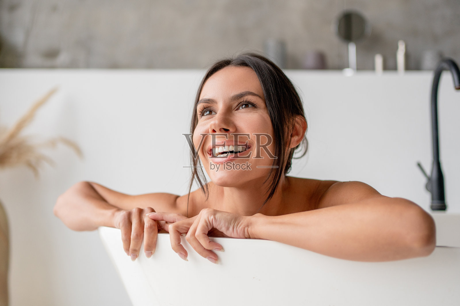 快乐的棕发女士坐在浴缸里洗澡,微笑着望向一旁。女性在浴室中享受沐浴时光。美容与健康的概念。照片摄影图片