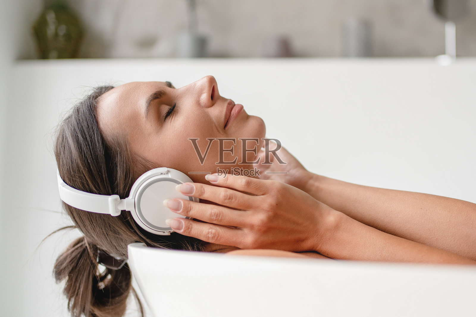 棕发白人女性在泡沫浴中闭眼聆听音乐,佩戴无线耳机,享受独自在家的周末,侧面特写肖像。照片摄影图片