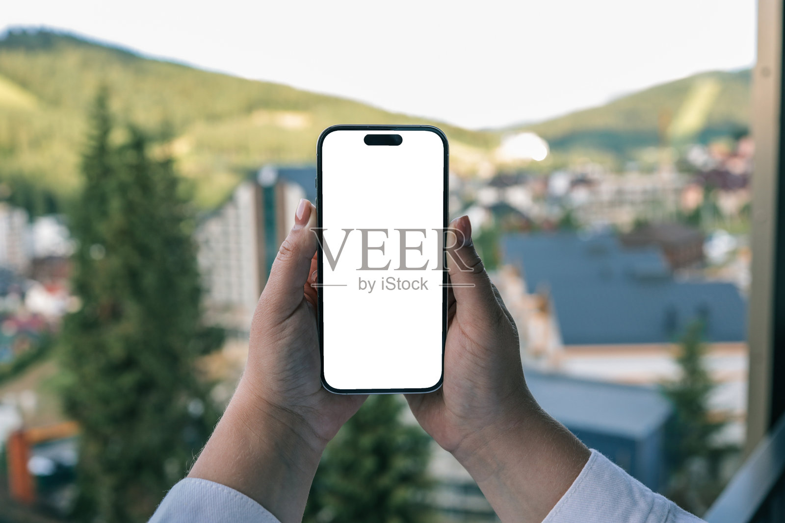 在阳台上拿着空白屏幕智能手机的人,背景模糊的房屋、屋顶和绿色山丘。科技、远程工作、数字通信、应用程序模型、模板的概念。照片摄影图片