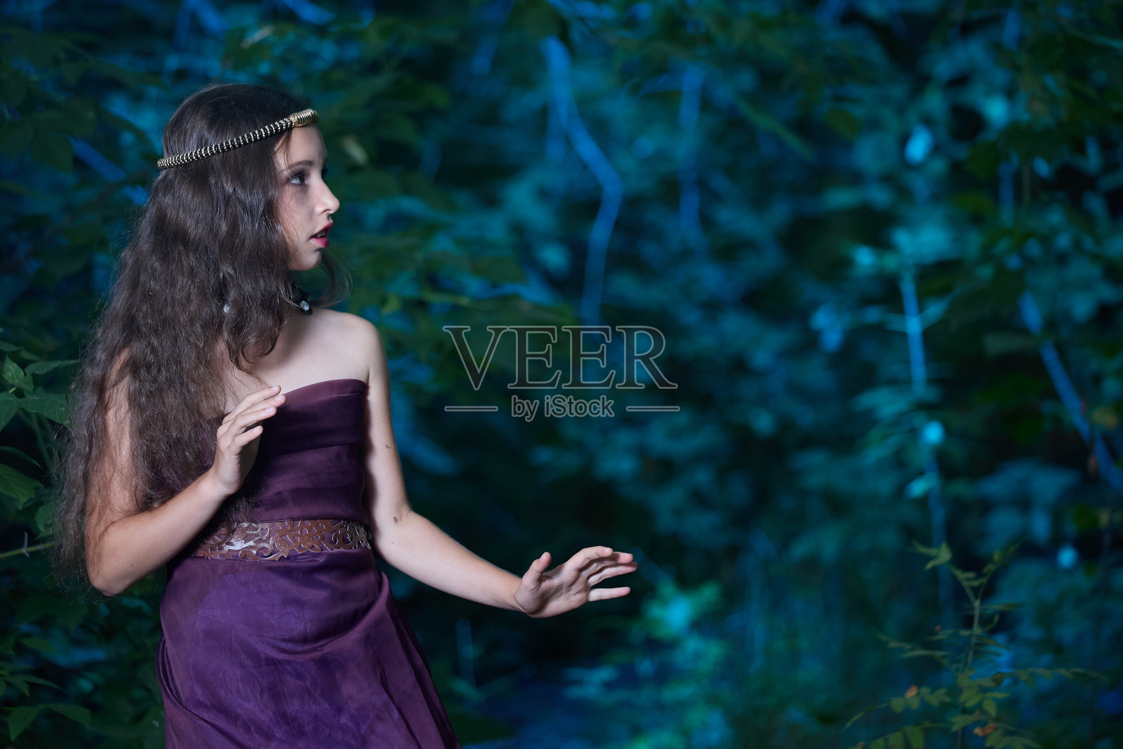 美丽的少女穿着长长的紫色裙子,站在森林里四处张望,神情害怕。照片摄影图片