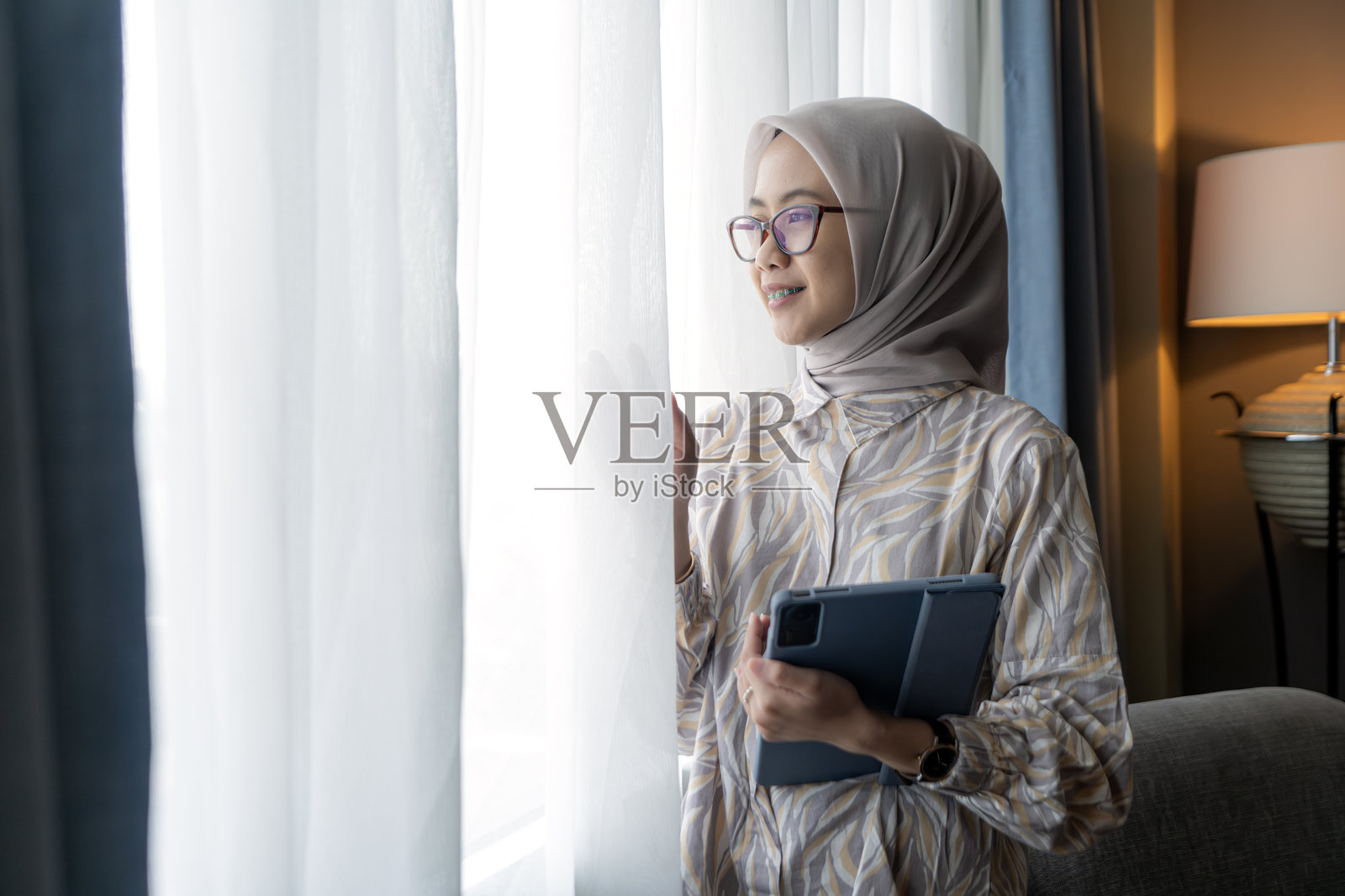 身穿头巾的印尼亚洲女性在室内透过窗户望出去,同时手持平板电脑。照片摄影图片