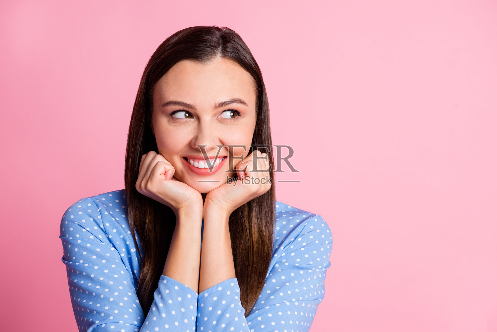 可爱的年轻女孩双手托腮,侧身望向空旷的空间,穿着蓝色波点衬衫,背景为柔和的粉色。照片摄影图片