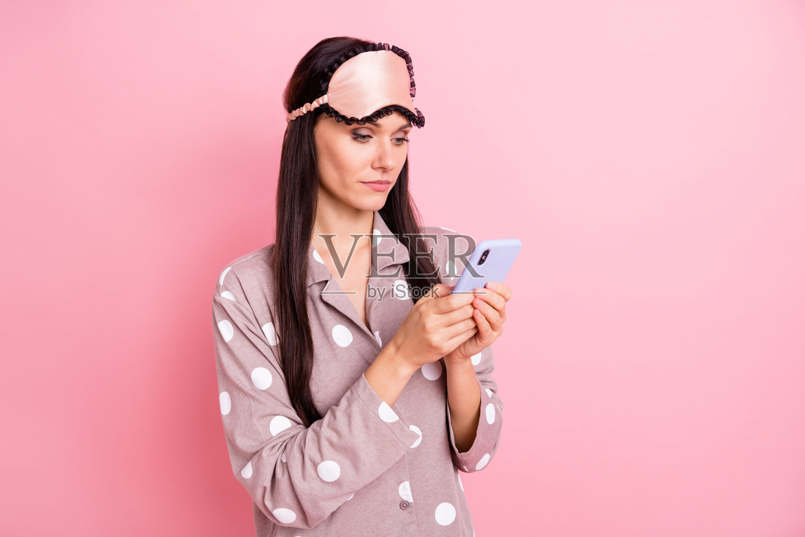 可爱的年轻女士穿着睡衣,戴着面罩,正在粉色背景下使用现代设备打字。照片摄影图片