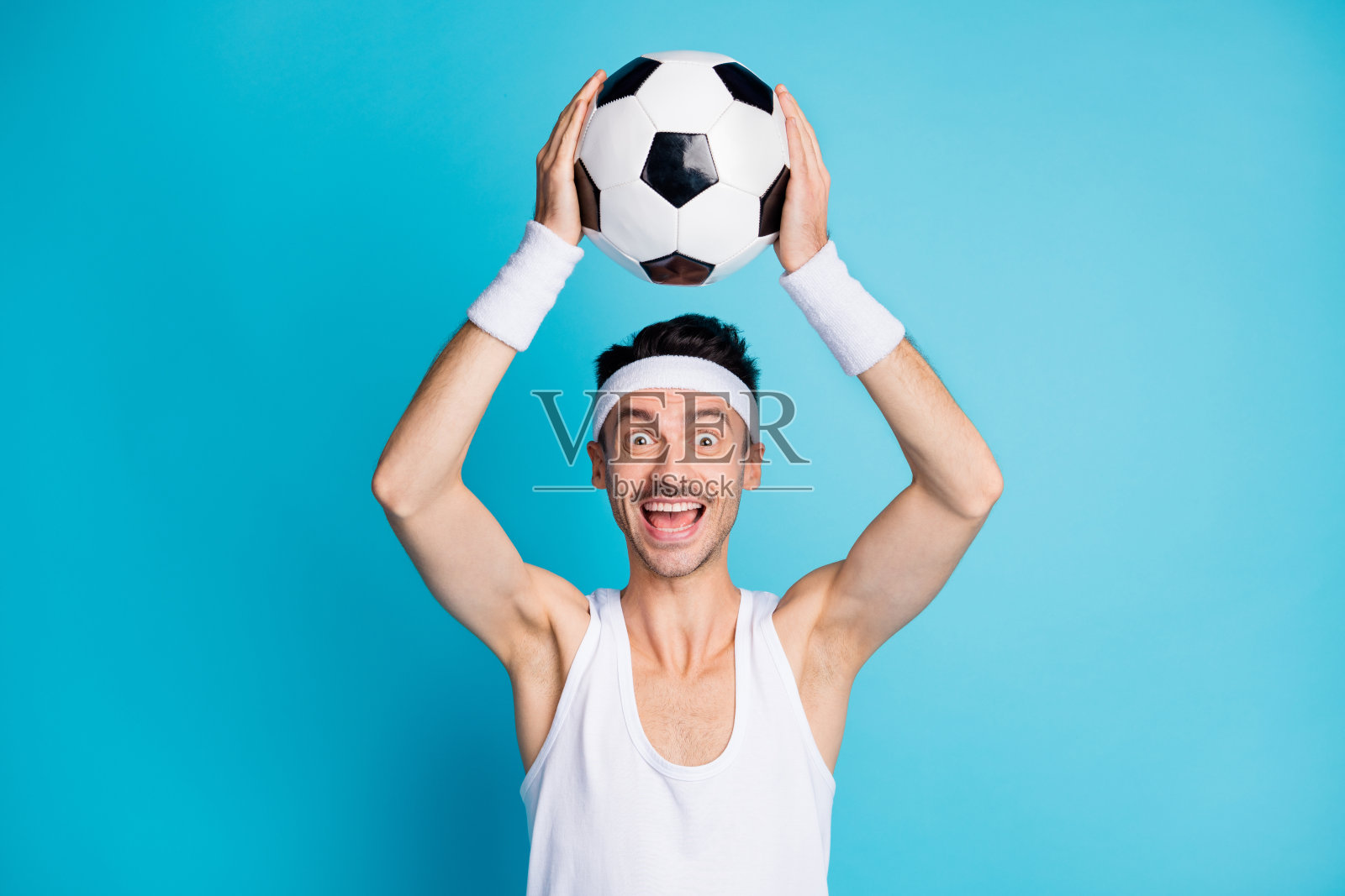 惊讶的足球运动员穿着白色背心,抓住足球并大喊,背景为蓝色。照片摄影图片