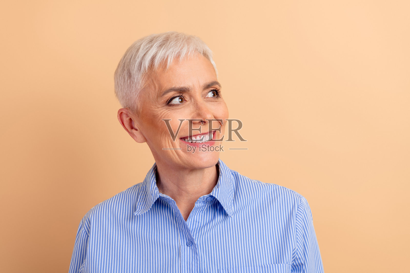 微笑的成熟的女性,短白发,穿着蓝色条纹衬衫,背景为米色,展现出积极和迷人的气质照片摄影图片