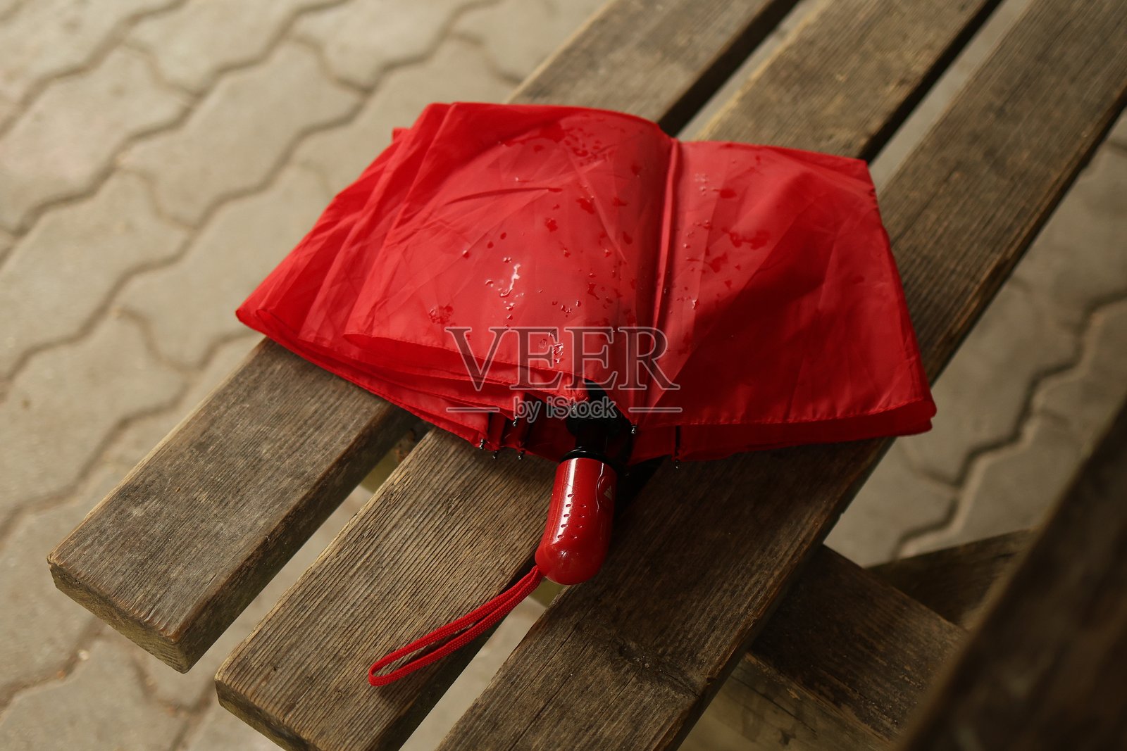 一把红色雨伞静静地放在户外的木长椅上。明亮的伞与自然的木材形成对比,营造出一种温馨而略带怀旧的氛围。照片摄影图片