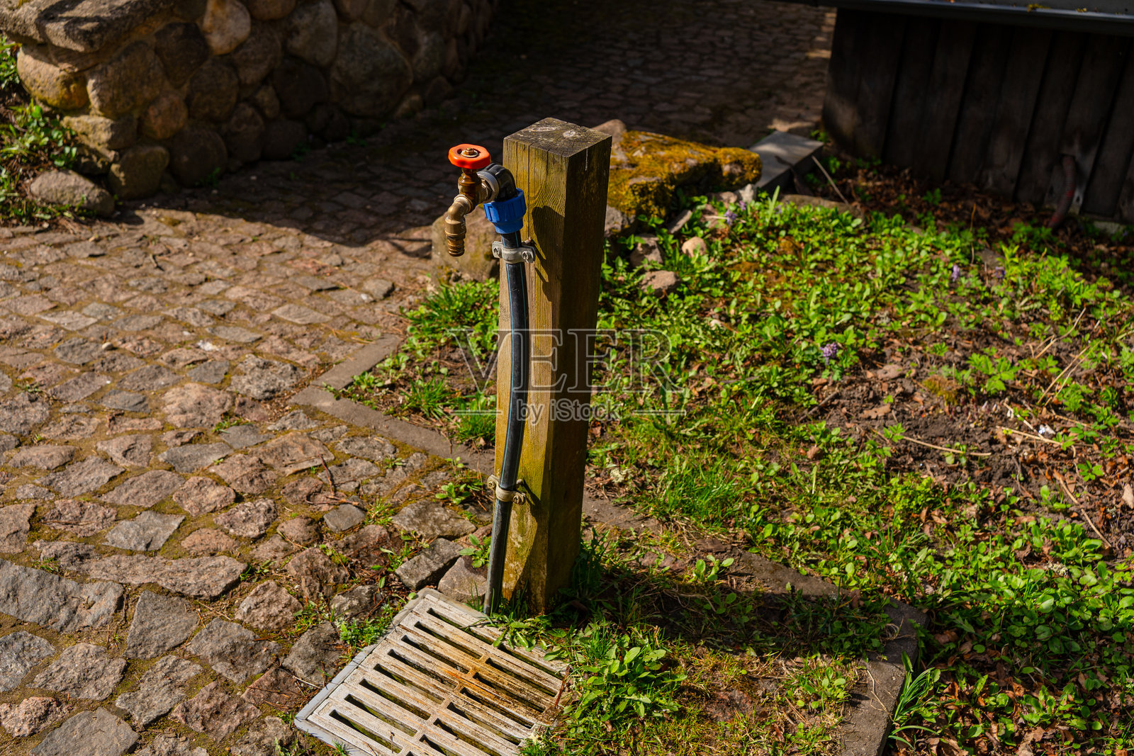 阳光照射下,后院花园中金属与塑料水龙头的特写,周围有木质支撑、灰色排水口和绿色植物。照片摄影图片