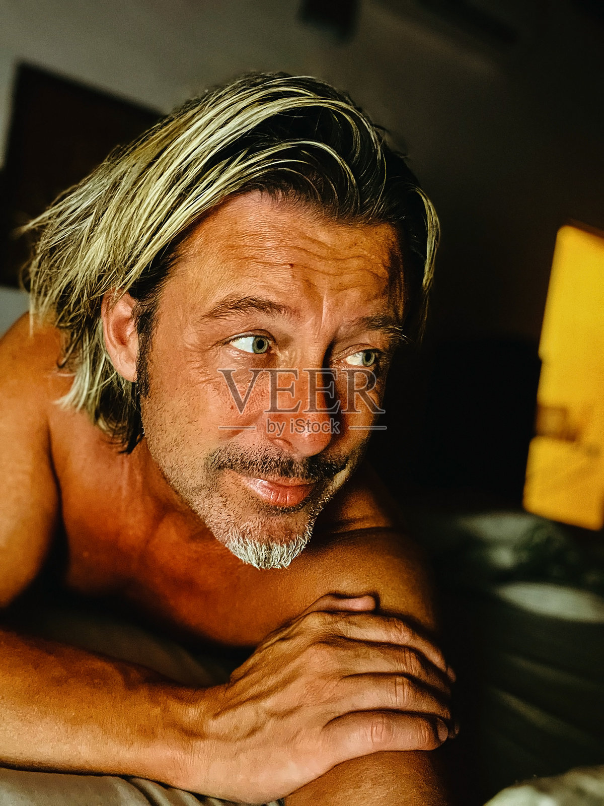 放松的晒黑金发男子,脸上有灰色胡茬,躺在卧室的床上,夜晚门开着。照片摄影图片