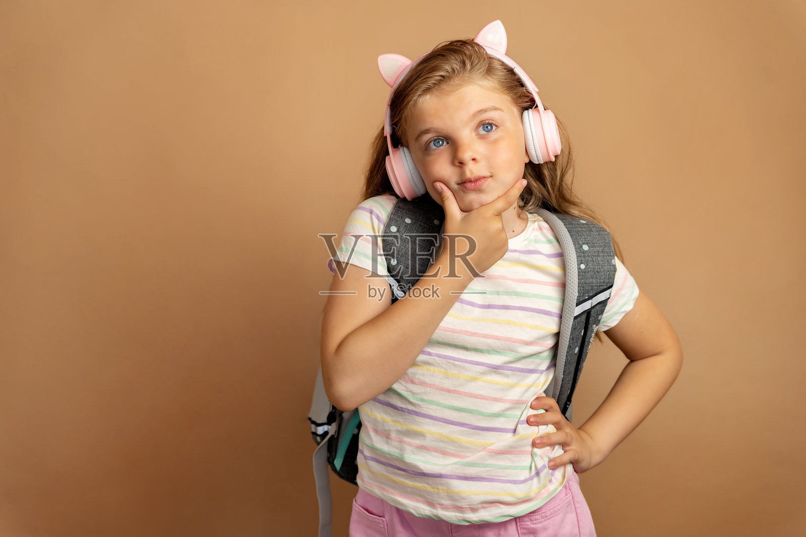 一个戴着耳机、背着书包的学龄儿童在米色背景下若有所思地望向一旁。在线教育。音乐与现代科技。照片摄影图片