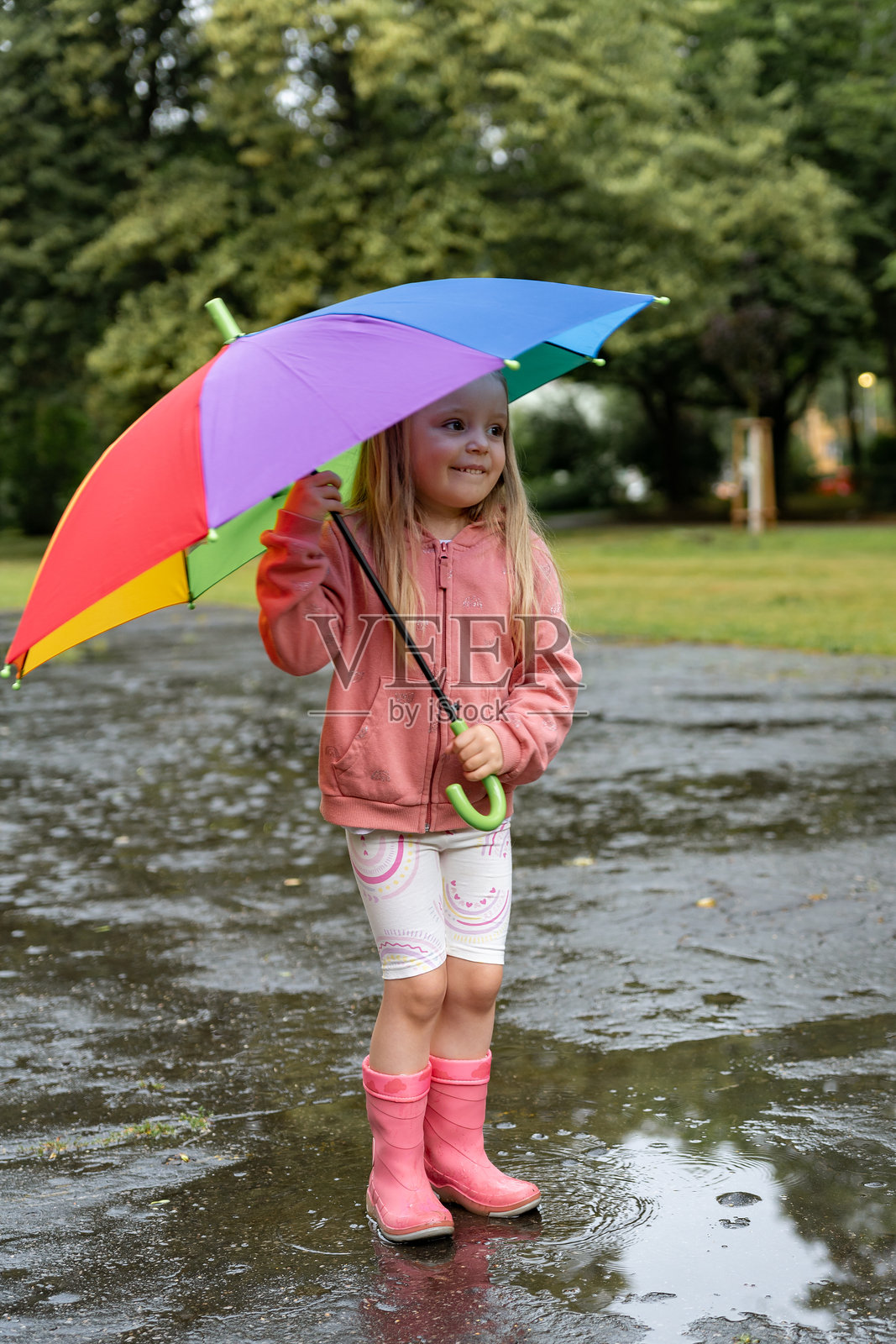 快乐的小女孩在夏天的雨中,手持五彩斑斓的雨伞站在水坑里。幼儿在公园的雨天中走过水坑。幸福的童年。照片摄影图片