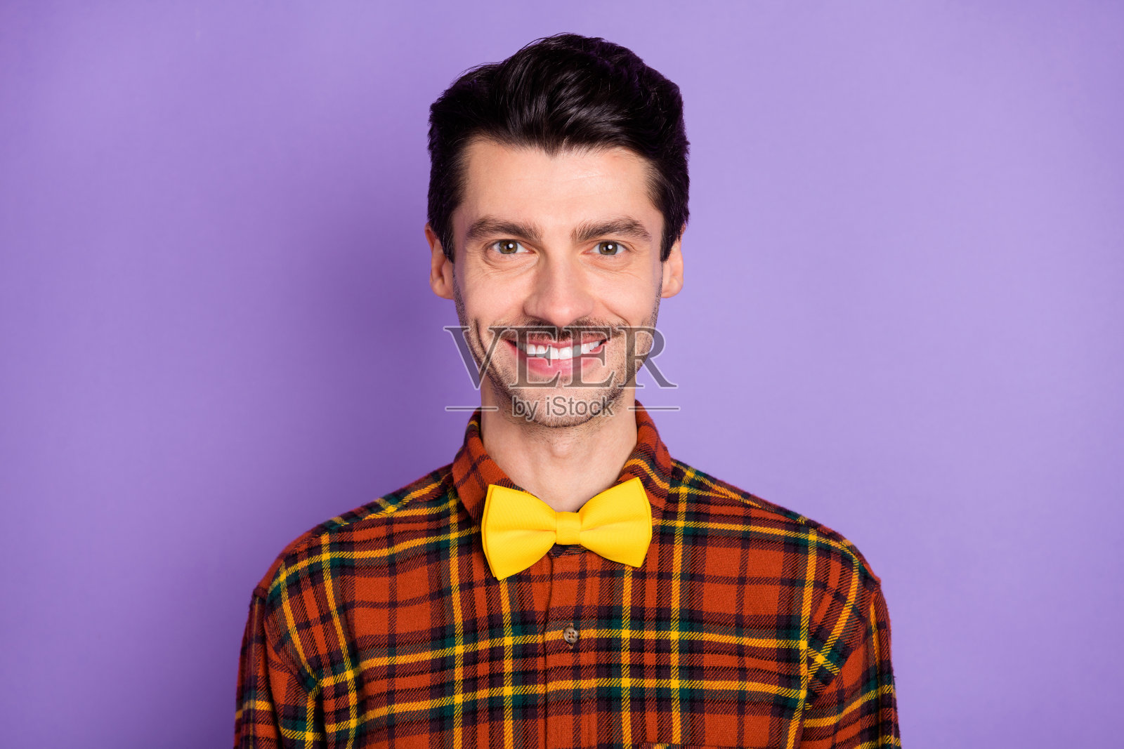 一位英俊年轻男子露出灿烂的笑容,穿着格子衬衫和蝴蝶结,面向镜头,背景为紫色。照片摄影图片