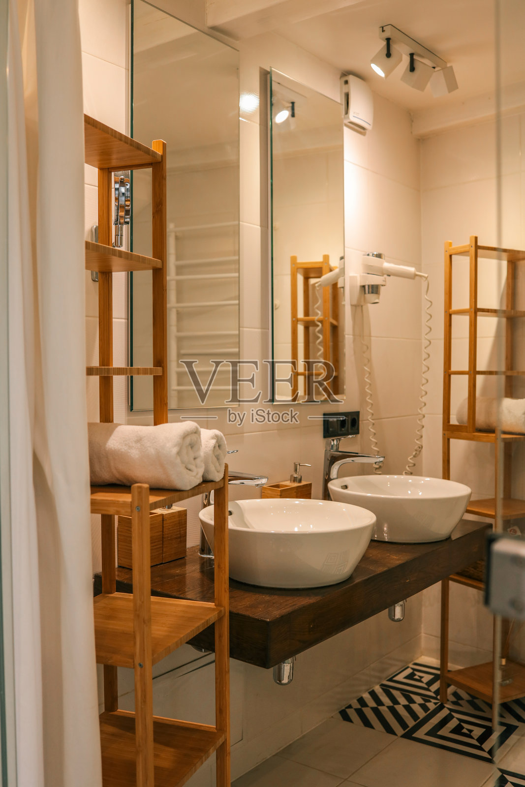 现代浴室内部,配有双白色陶瓷洗手盆、木质架子和卷起的毛巾,特色图案瓷砖和温暖的灯光。照片摄影图片