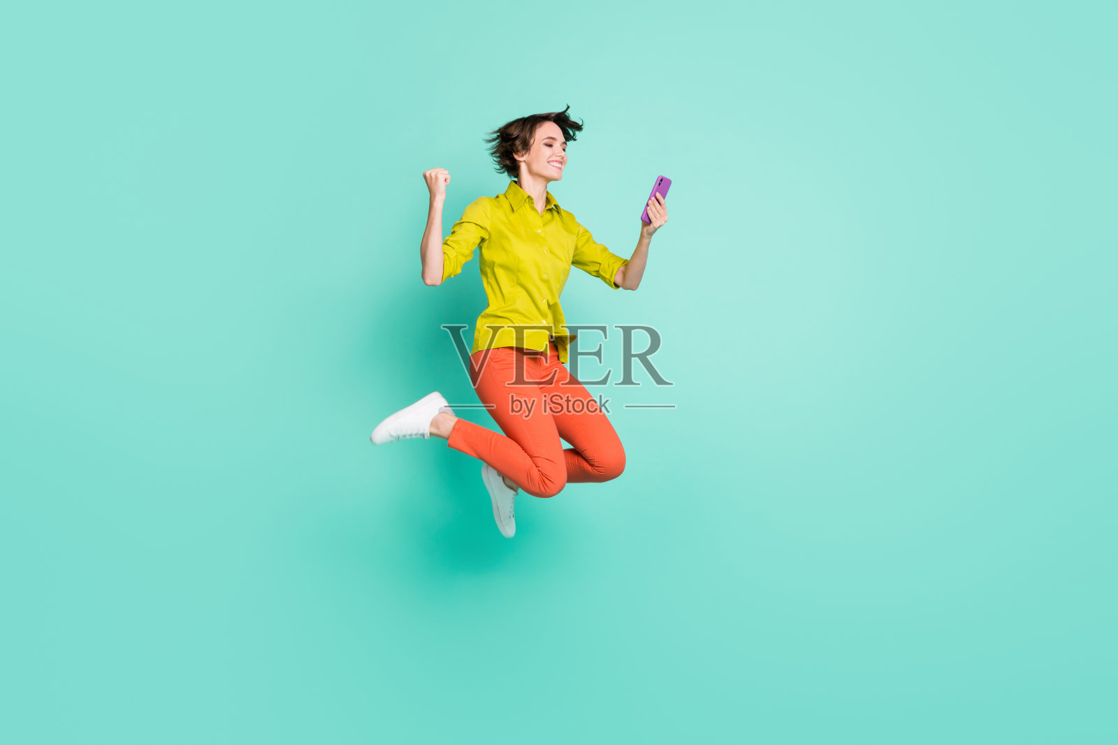 高高跳起的女性全身照,手持智能手机,做出胜利手势,背景为鲜艳的青色。照片摄影图片