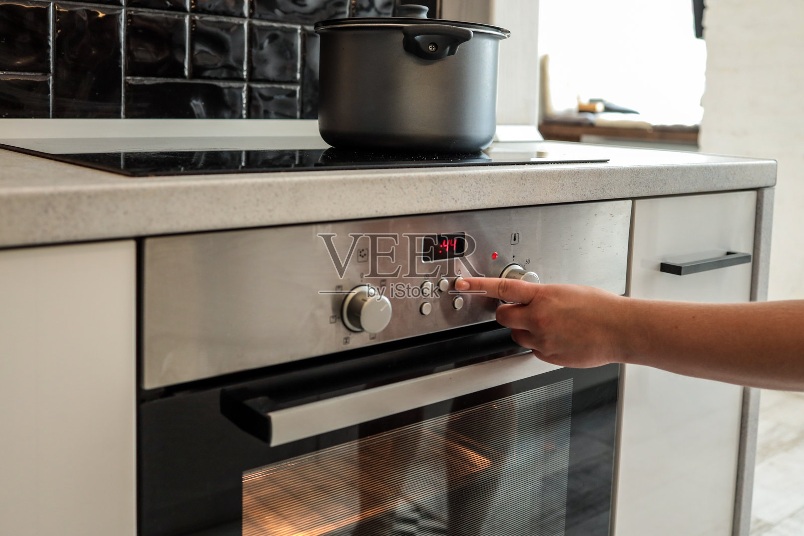 一个人正在使用控制按钮设置现代内置烤箱的时间或温度。烤箱正在工作,发出光芒,上面放着一个黑色锅。照片摄影图片