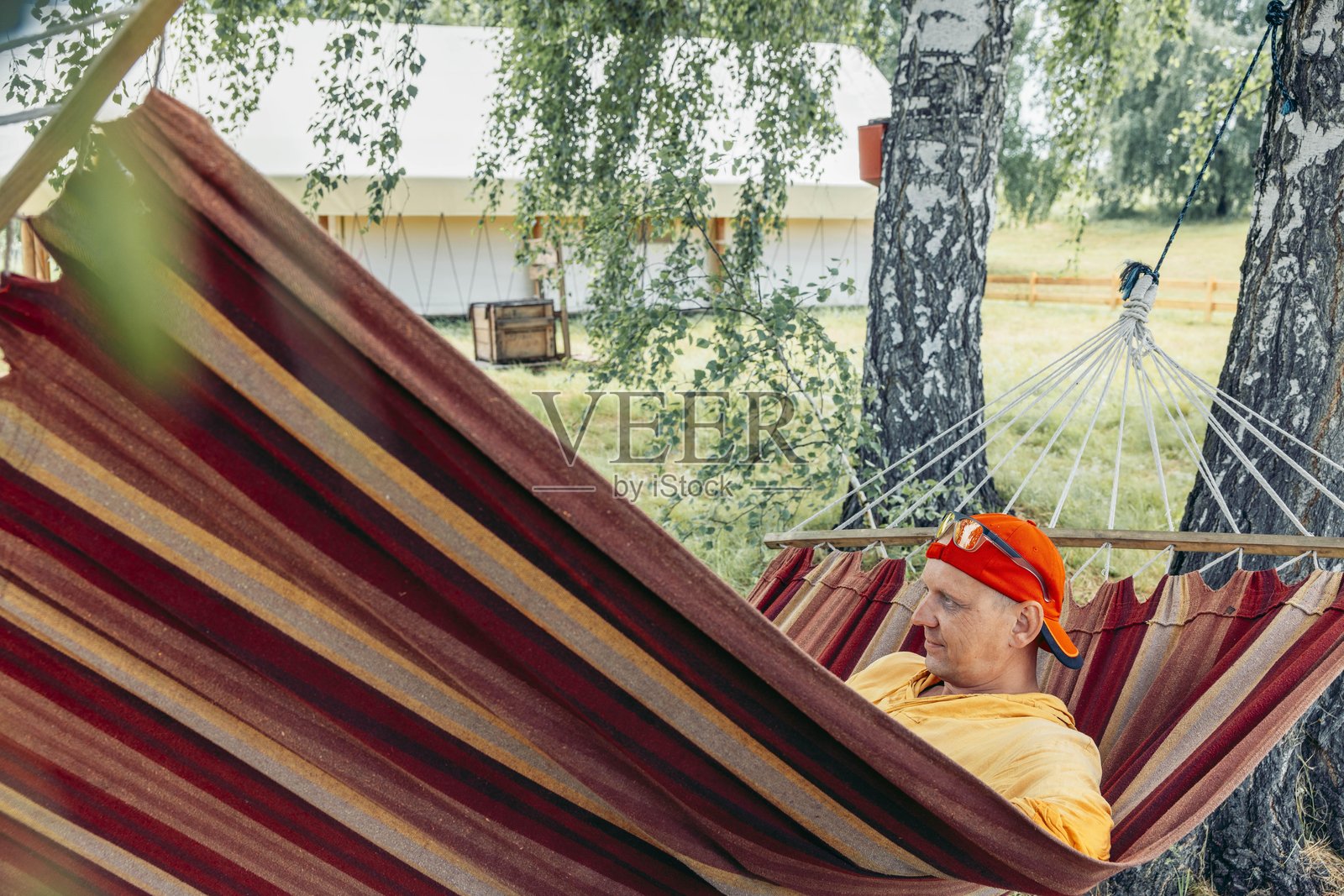 夏季露营度假中,戴着红色帽子、穿着黄色卫衣的男子在树下的条纹吊床上放松,背景是豪华露营帐篷,体现了户外休闲旅游度假营销的概念。照片摄影图片