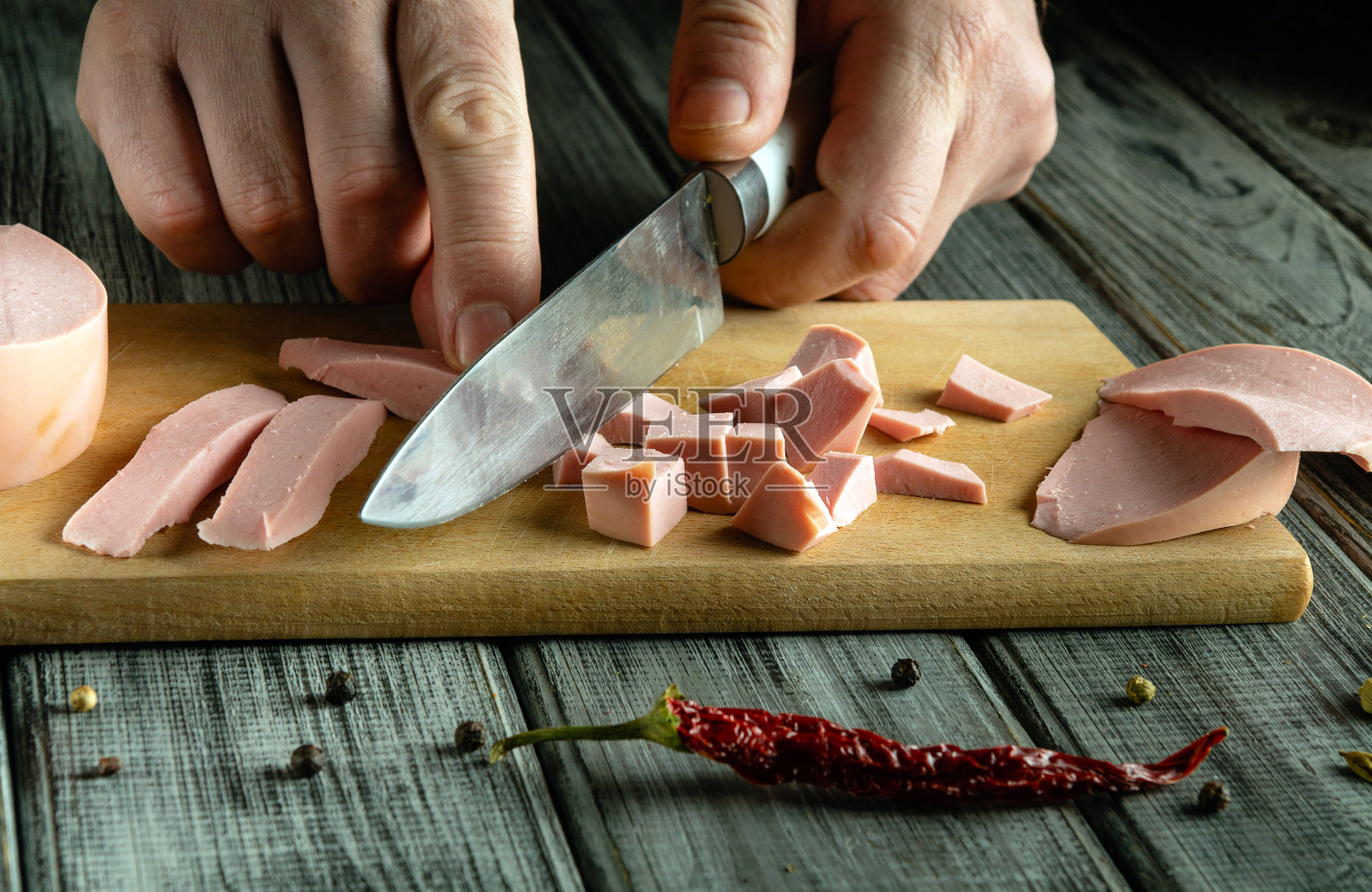 熟练的手将冷切肉切成小方块,放在木板上。干辣椒和整颗黑胡椒散落在质朴的木桌上。照片摄影图片
