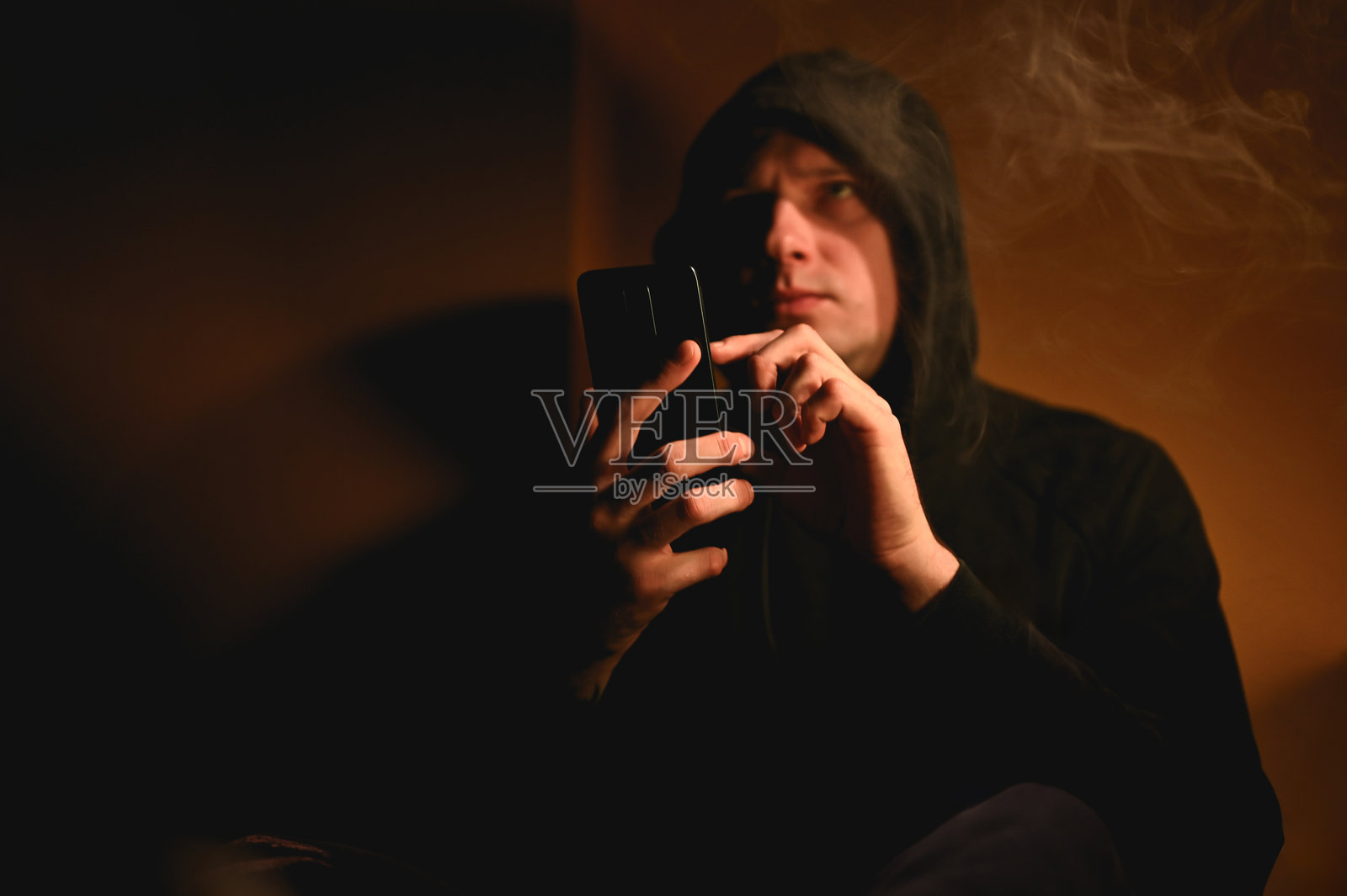 在烟雾弥漫的房间里,戴着兜帽的男子正在浏览智能手机照片摄影图片