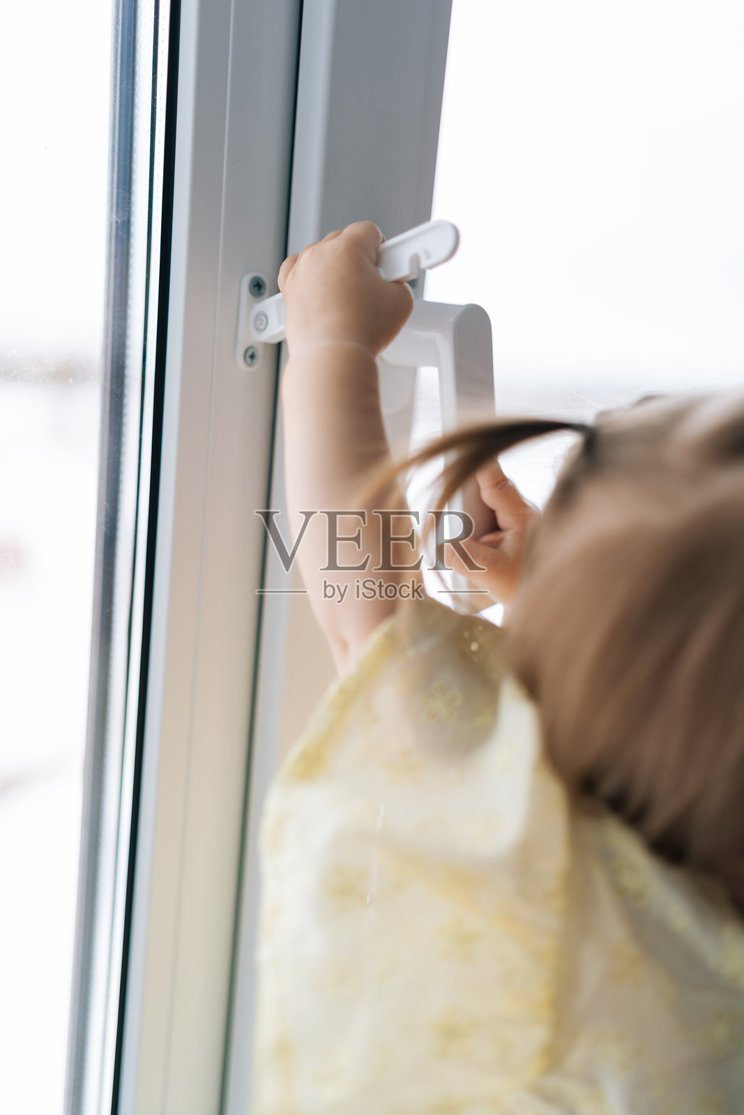 好奇的小女孩竖直裁剪的照片,手伸向窗户把手。潜在家庭安全风险的概念。照片摄影图片