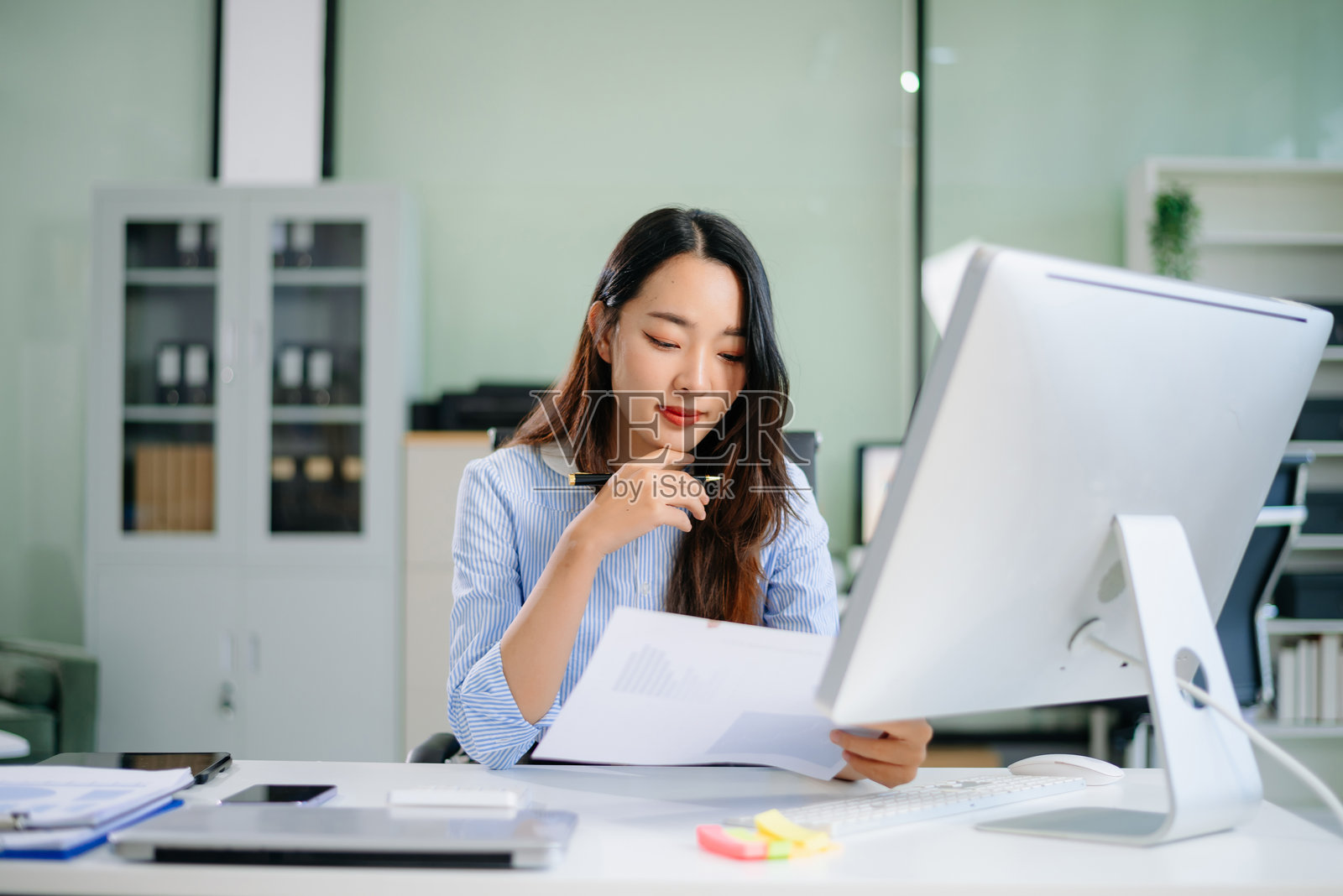 亚洲女商人在明亮现代的办公室里站在桌子旁,手持夹板写字,同时分析数据图表。照片摄影图片