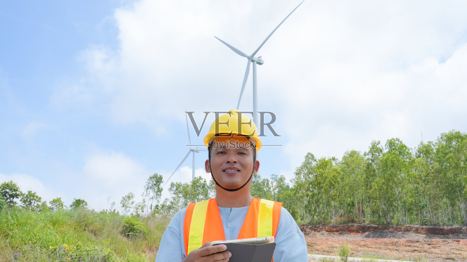 微笑的工程师站在风力涡轮机前,戴着安全帽和安全背心,手持平板电脑。场景传达出可再生能源中的进步与可持续性的感觉。照片摄影图片