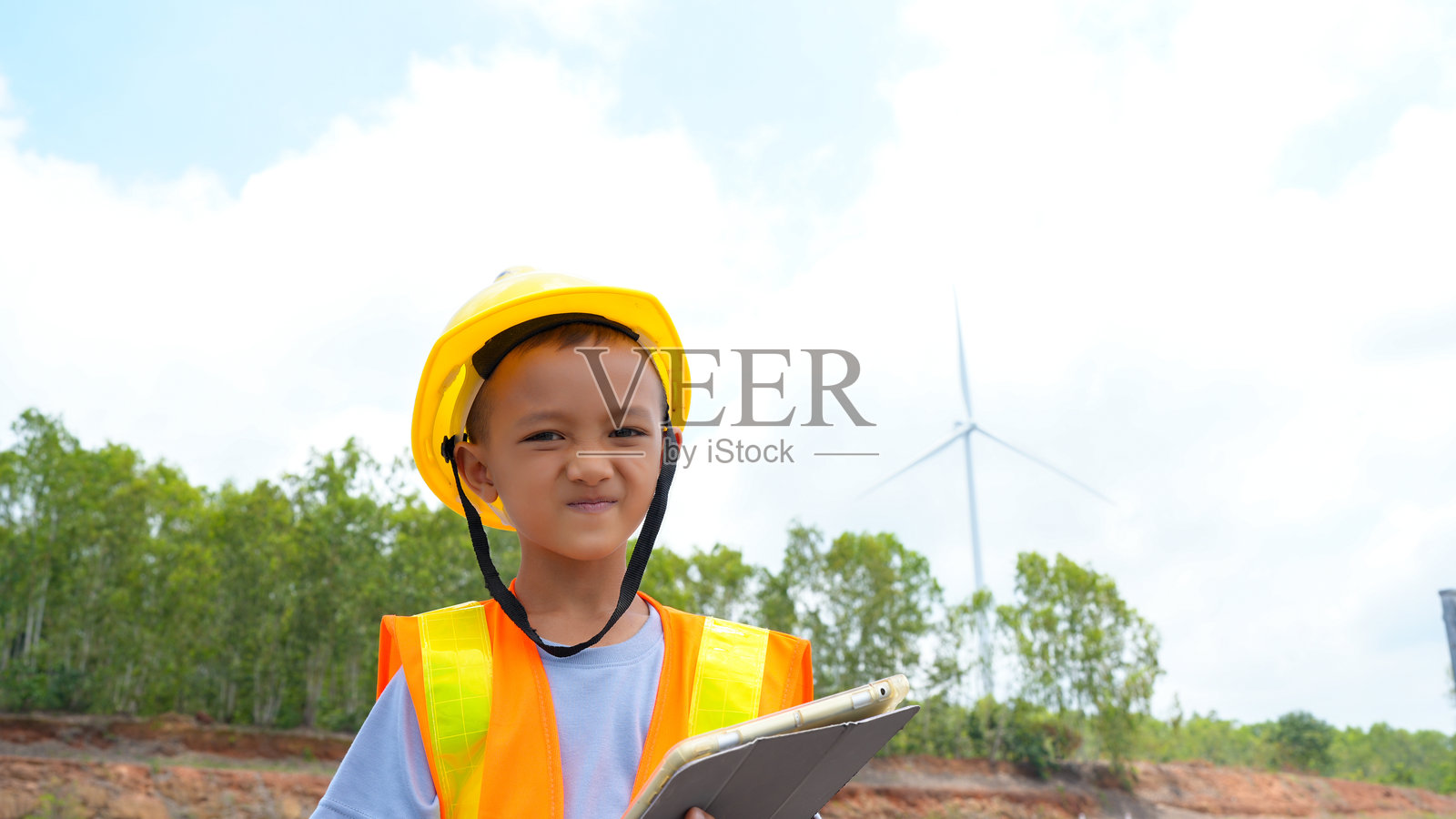 年轻儿童佩戴安全头盔和反光背心,自信地站在建筑工地上,手持平板电脑。背景中可见风力发电机,象征着可再生能源和创新。照片摄影图片