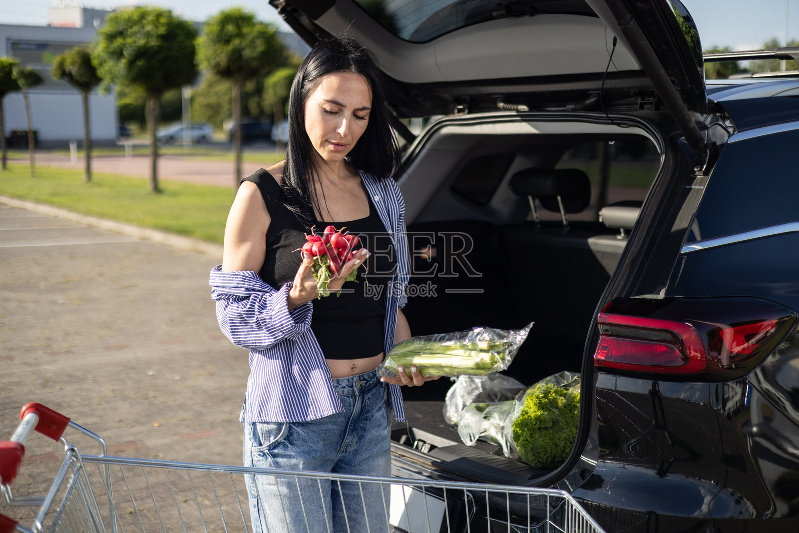 年轻的棕发女性站在超市停车场,旁边是一辆打开后备箱的车,她正在将购物车里的杂货转移到车的后备箱中。照片摄影图片