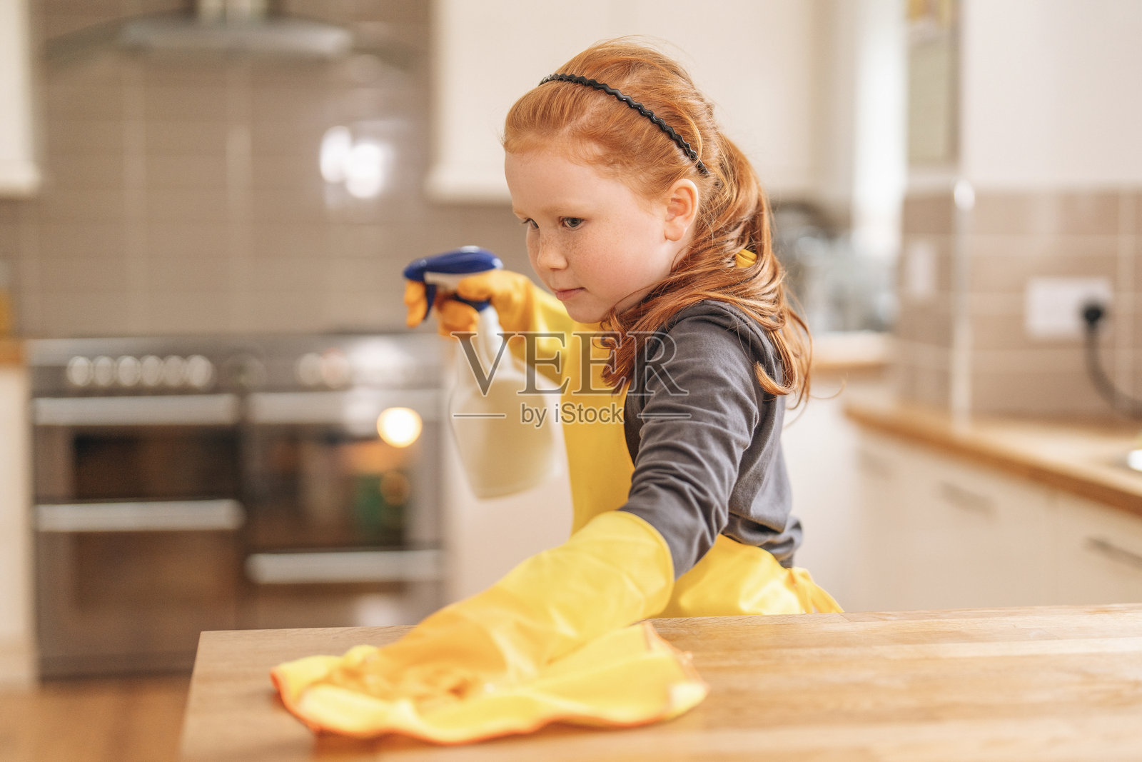 年轻女孩在明亮的白天穿着黄色手套和围裙,使用喷雾瓶清洁厨房台面。照片摄影图片