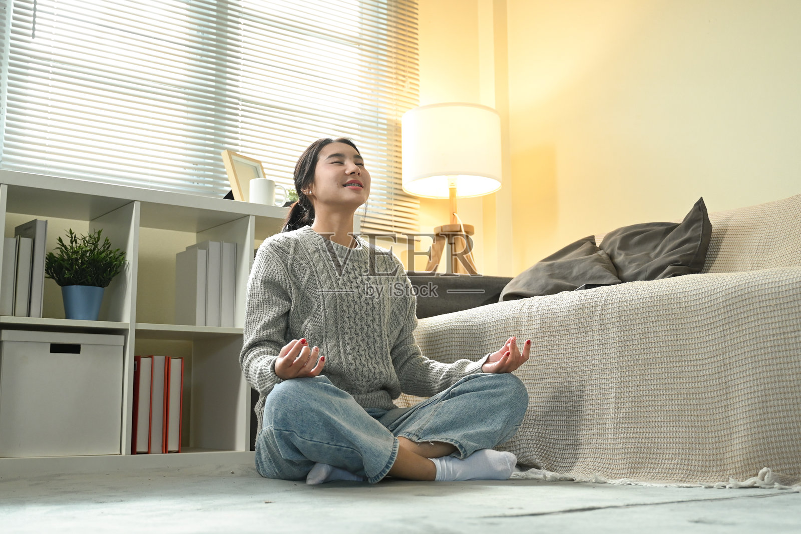 一位女性坐在客厅的地板上,穿着灰色毛衣和蓝色牛仔裤。她处于放松状态,可能在冥想或练习瑜伽。照片摄影图片
